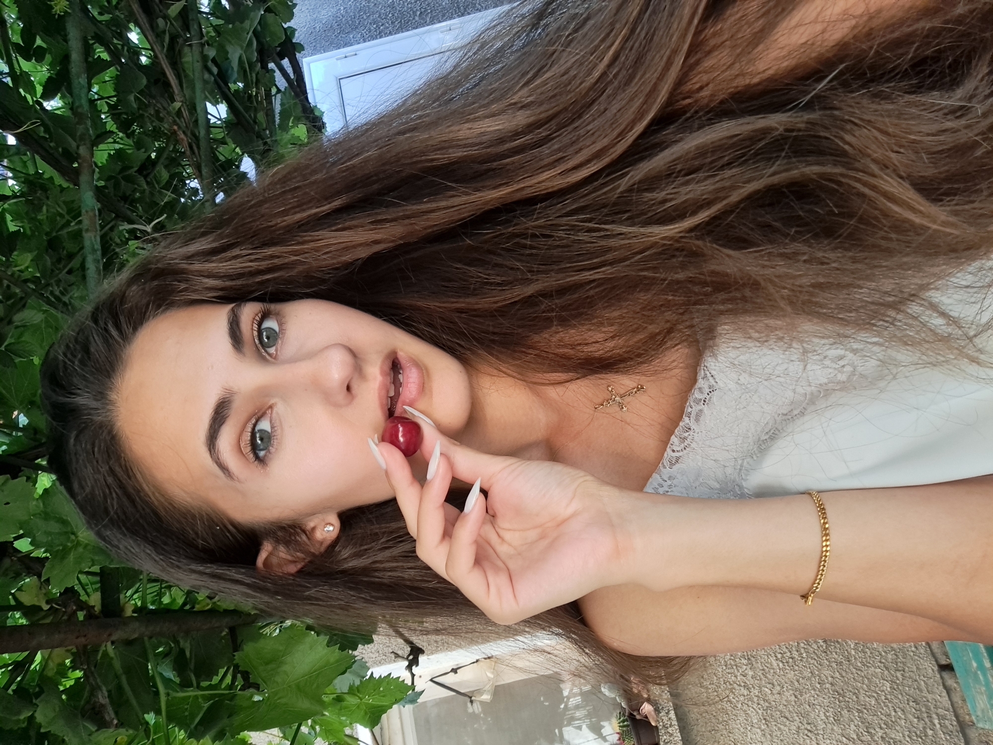 A young woman with long, dark brown hair and blue-green eyes is shown from the chest up, holding a single cherry near her lips. She is wearing a white lace-trimmed top and a gold bracelet on her left wrist. A gold cross necklace is visible around her neck. The background suggests an outdoor setting with green foliage and a textured wall, possibly a building exterior. The lighting indicates it is daytime, with the sun illuminating her face and jewelry. The image appears to be a selfie taken outdoors in a residential or garden area. Based on its geographical coordinates, the image was taken in the village of Chepintsi, Bulgaria.