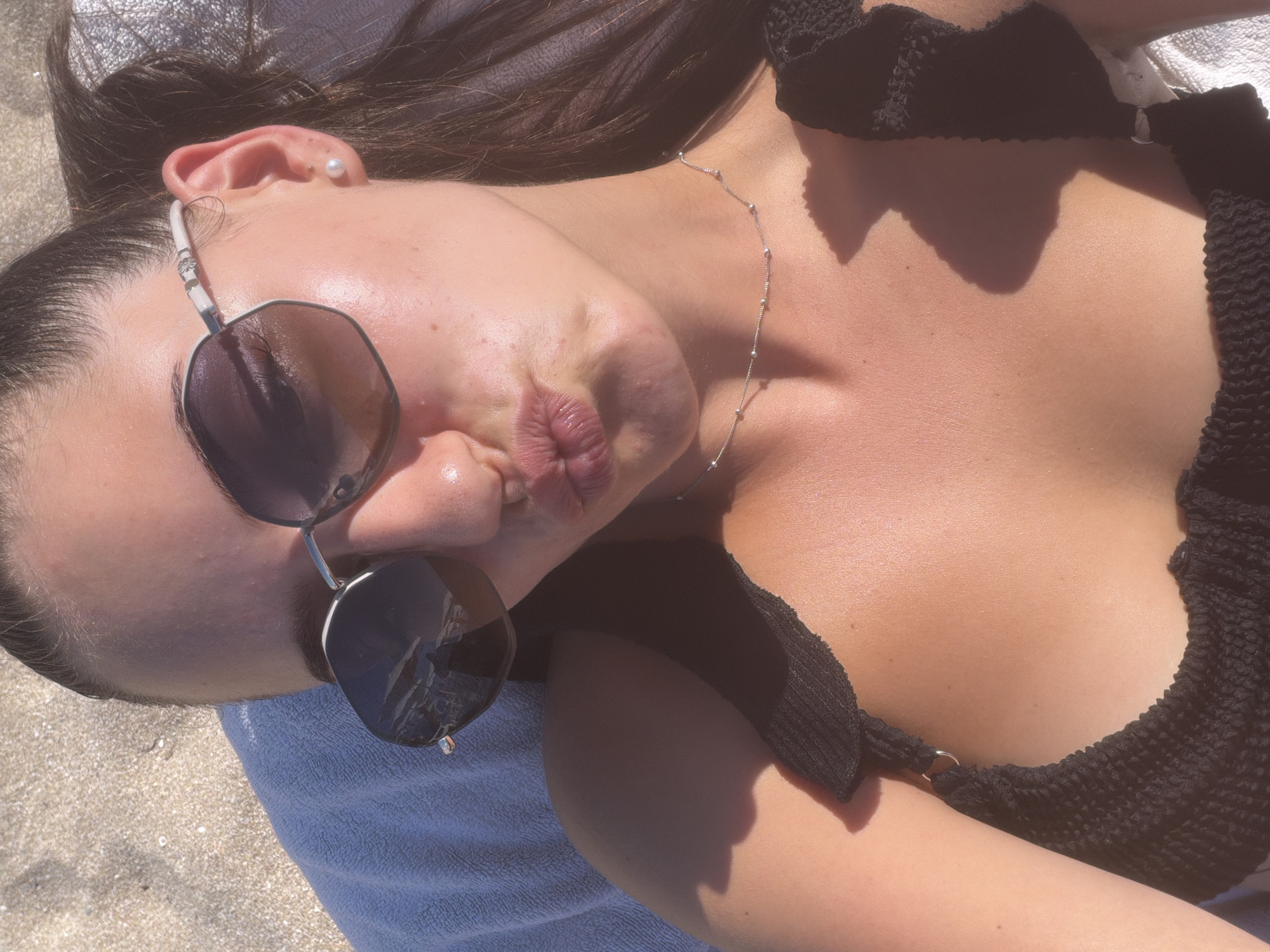 In the city of Lozenets, Bulgaria, a woman with dark hair pulled back is lying on a beach towel on the sand. She is wearing a black bikini top, sunglasses, and a delicate silver necklace. The woman is taking a selfie, puckering her lips towards the camera. The sunlight casts shadows on her skin and the surrounding sand.