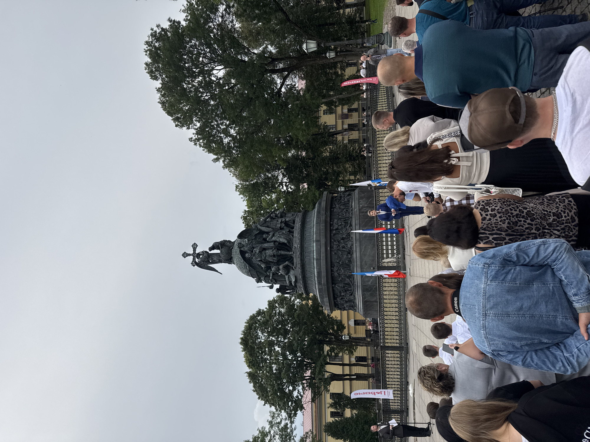The image captures an outdoor gathering in front of the Millennium of Russia monument in Veliky Novgorod, Russia. A crowd of approximately thirty people is visible, facing a monument topped with an angel and surrounded by historical figures. Two men are speaking from a raised platform, one in a dark suit and the other in a blue jacket, addressing the assembly. Several flags, including Russian flags, are displayed near the monument. The environment appears to be a public square with paved ground, a decorative fence, and mature trees. The sky is overcast, suggesting a cloudy day. The overall scene depicts a formal event or ceremony taking place at a prominent historical landmark.