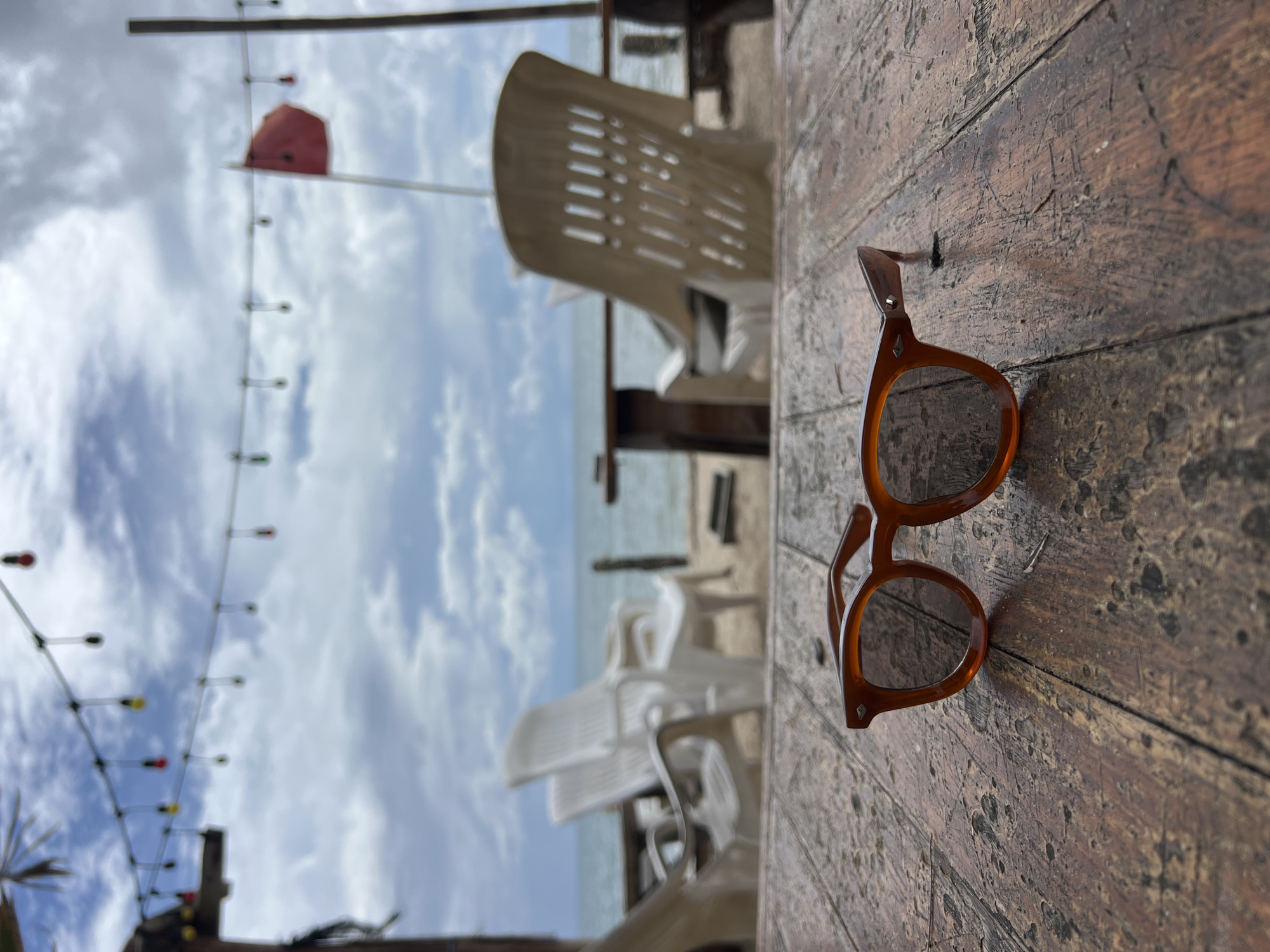 An outdoor scene at a beach resort features a pair of brown-rimmed sunglasses on a weathered wooden table in the foreground. In the middle ground, two white plastic lounge chairs are positioned on a sandy area facing the ocean. Further back, a red flag flies from a pole, with a string of colorful lights overhead against a cloudy sky. The ocean stretches to the horizon, with a calm sea and overcast weather.