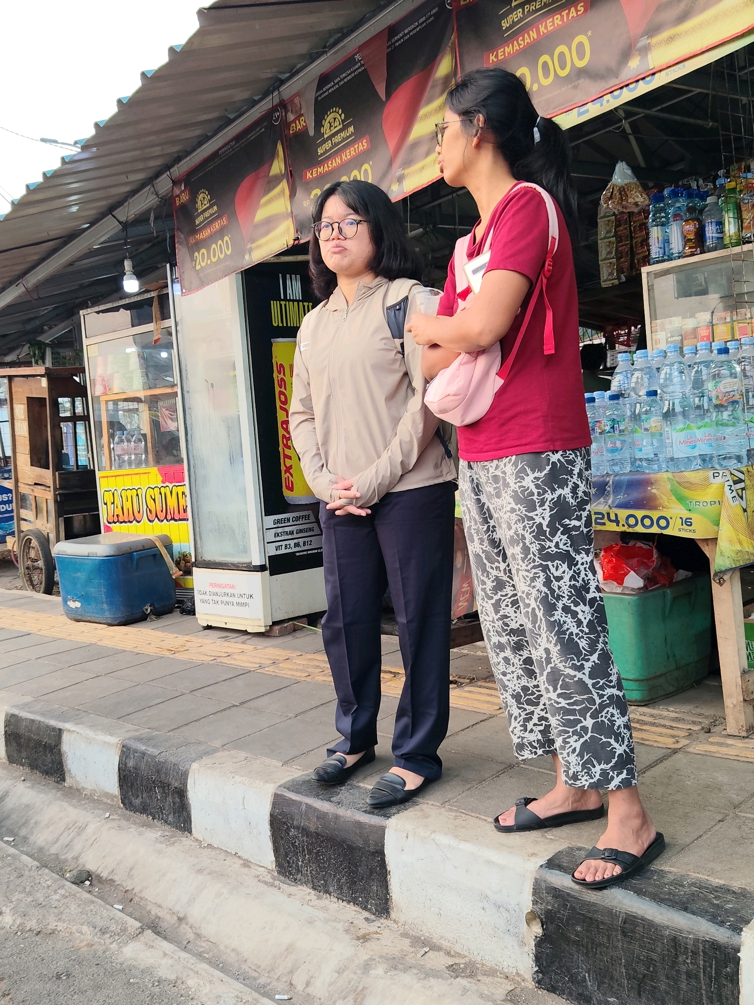Two women stand on a sidewalk next to a street in Cikampek, Indonesia. The woman on the left wears glasses, a light brown jacket, and dark pants. The woman on the right has dark hair pulled into a ponytail, wears a red shirt, and patterned pants. She also has a pink bag slung over her shoulder. They are positioned in front of a stall or shop with awnings overhead. The stall displays various bottled drinks and some food items. A blue cooler sits on the sidewalk near the stall, and a cart with a wheel is visible to the left. The sidewalk has a curb with black and white stripes. The ground is paved, and a concrete drain runs alongside the street.