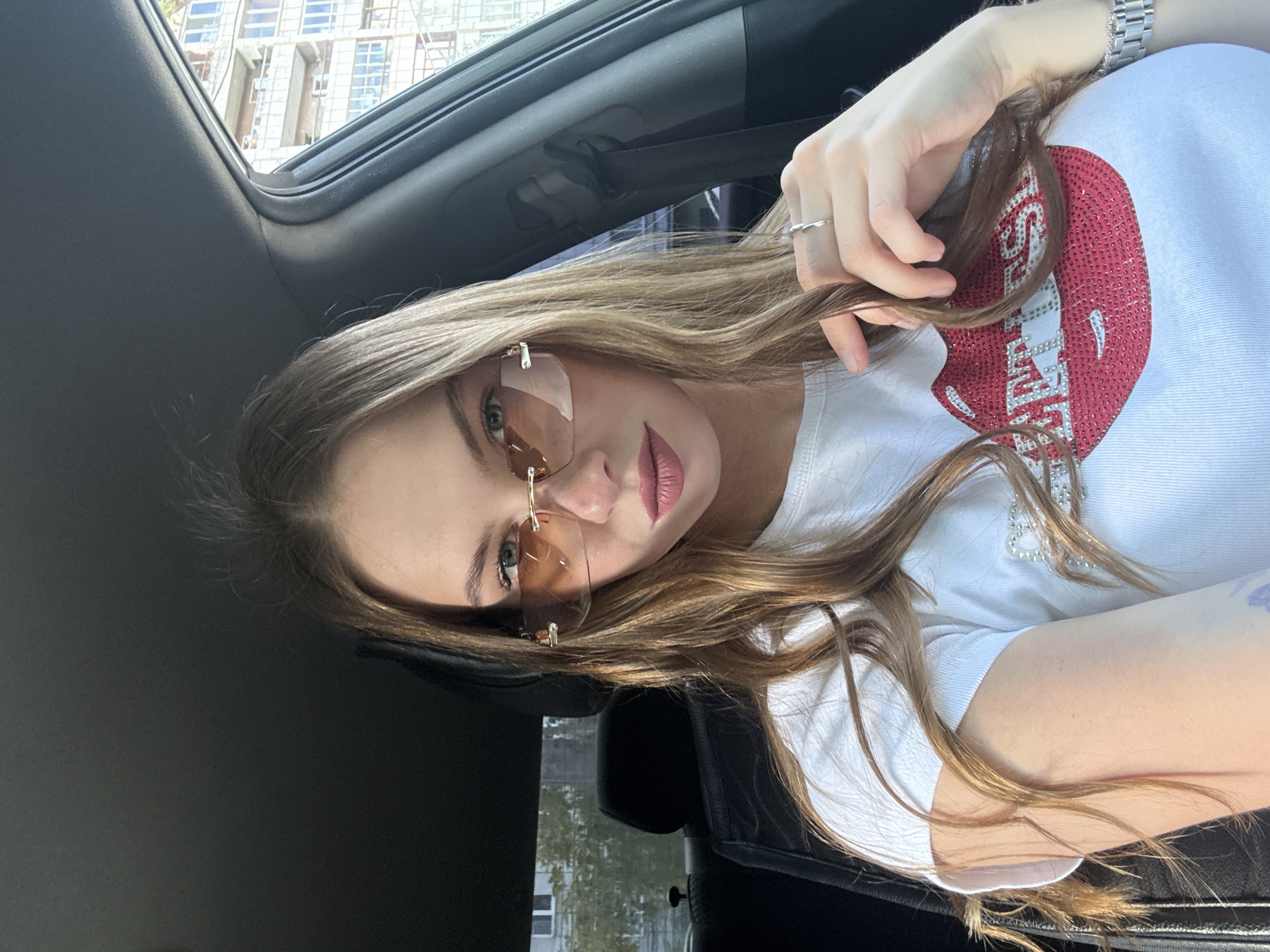 A young woman with long blonde hair is sitting in the driver's seat of a car, facing forward and looking at the camera. She is wearing rimless sunglasses with amber lenses and a white t-shirt with a large red lip graphic adorned with rhinestones. Her left hand is resting on her hair, and she is wearing a silver ring on her index finger. A silver watch is visible on her left wrist. The interior of the car, including the headrest and part of the door panel, is visible. Outside the car window, a building with multiple floors and windows can be seen. The image appears to be taken during the daytime, with sunlight illuminating the scene. The building and surroundings suggest an urban environment, consistent with the city of Perm, Russia.