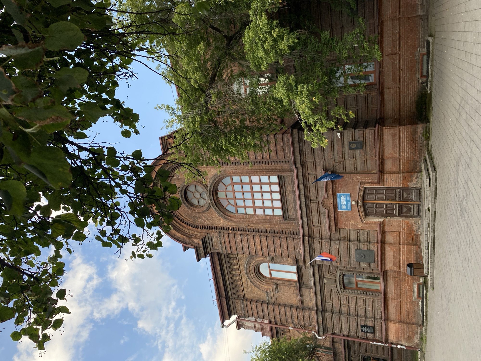 The image shows a large, old, brick building with multiple arched windows and a prominent arched entryway. A Russian flag is displayed on the facade to the left of the entrance. There are trees with green foliage partially obscuring the building, suggesting a temperate climate and possibly spring or summer. The building appears to be a public institution, as indicated by a blue sign above the entrance and what look like plaques on the walls. The sky is partly cloudy with a mix of blue and white. The foreground is a paved area, likely a sidewalk or courtyard. This building is the Krasnoyarsk State Pedagogical University named after V.P. Astafyev.