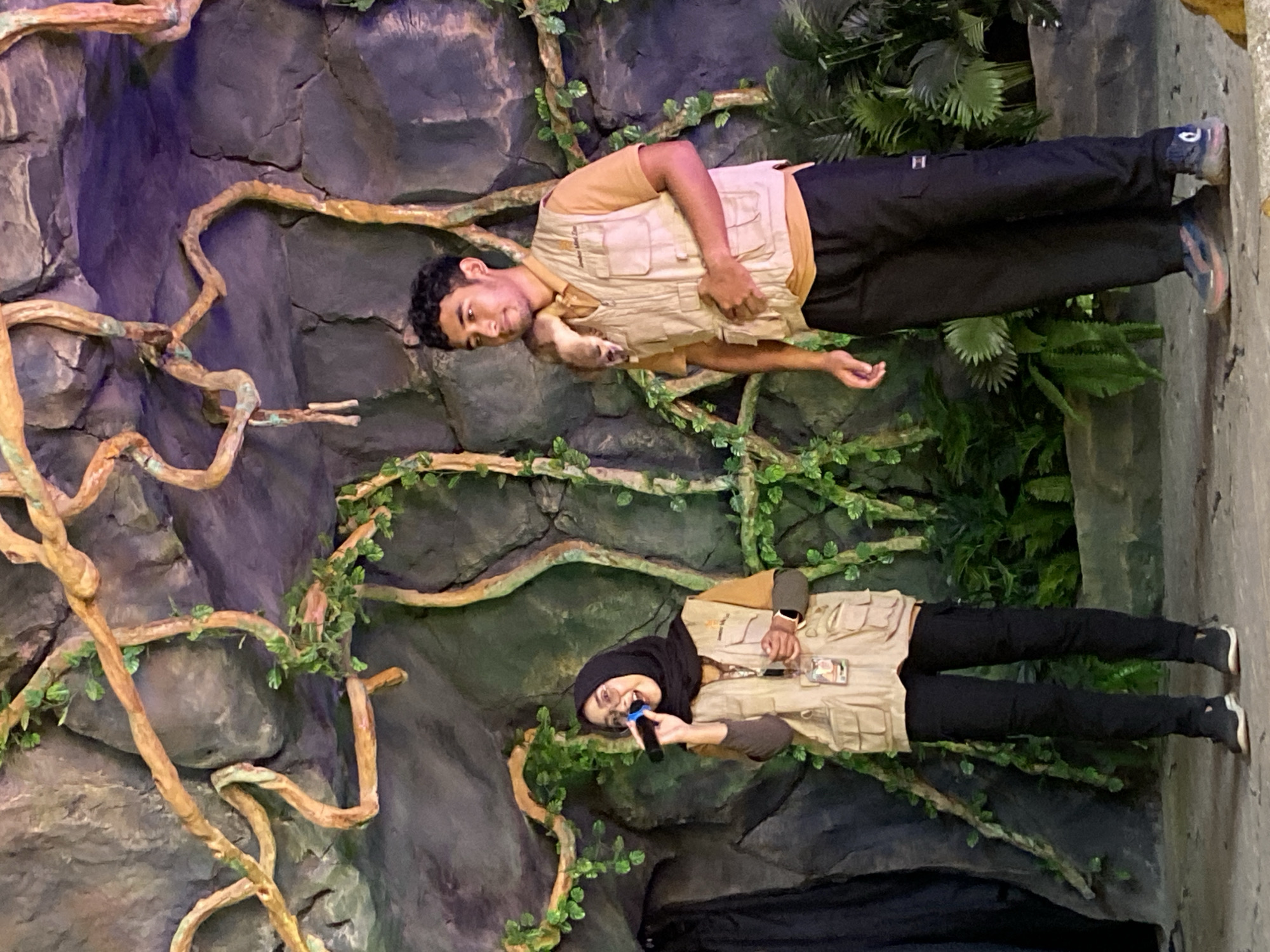 The image captures two individuals in what appears to be a themed environment, possibly a zoo or wildlife park in Mangli, Indonesia. The background is a faux rock wall adorned with artificial vines and greenery, simulating a jungle or rainforest setting.
The individuals, a man and a woman, are both wearing uniforms that suggest they are staff members. The man is wearing a beige vest over a tan shirt and dark pants, while holding a small, presumably live, animal, possibly a mammal. The woman, wearing a black hijab, is dressed in a similar vest over a brown shirt and dark pants, and is holding a microphone.
The scene suggests an educational presentation or demonstration about wildlife.