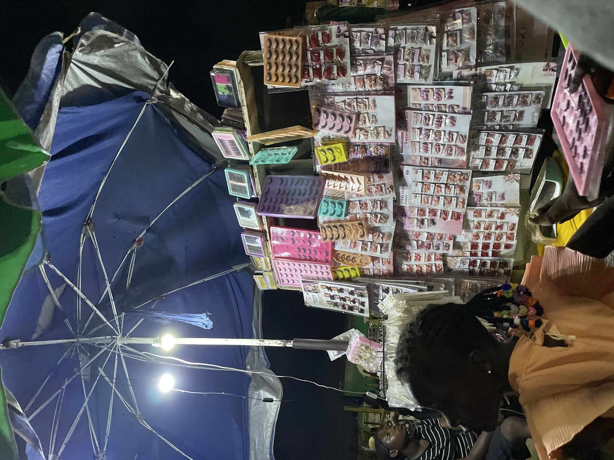In Lekki, Nigeria, at night, a stall is set up under a large blue and green umbrella. Two bright lights illuminate the stall, which displays a variety of artificial eyelashes and fingernails. These beauty products are neatly arranged on shelves and hanging displays, some in colorful packaging.
In the foreground, a young Black girl with braided hair decorated with colorful beads is seated and looking to her left. She wears an orange ruffled top. To her left, another person, possibly a young Black woman, is seated and wearing a striped shirt. Their legs are visible in the lower portion of the image. One person's feet are resting near a small green tray, and another person is holding a pink container filled with artificial eyelashes. The background is dark, indicating it is nighttime. The overall scene suggests a bustling night market or street vendor setup.