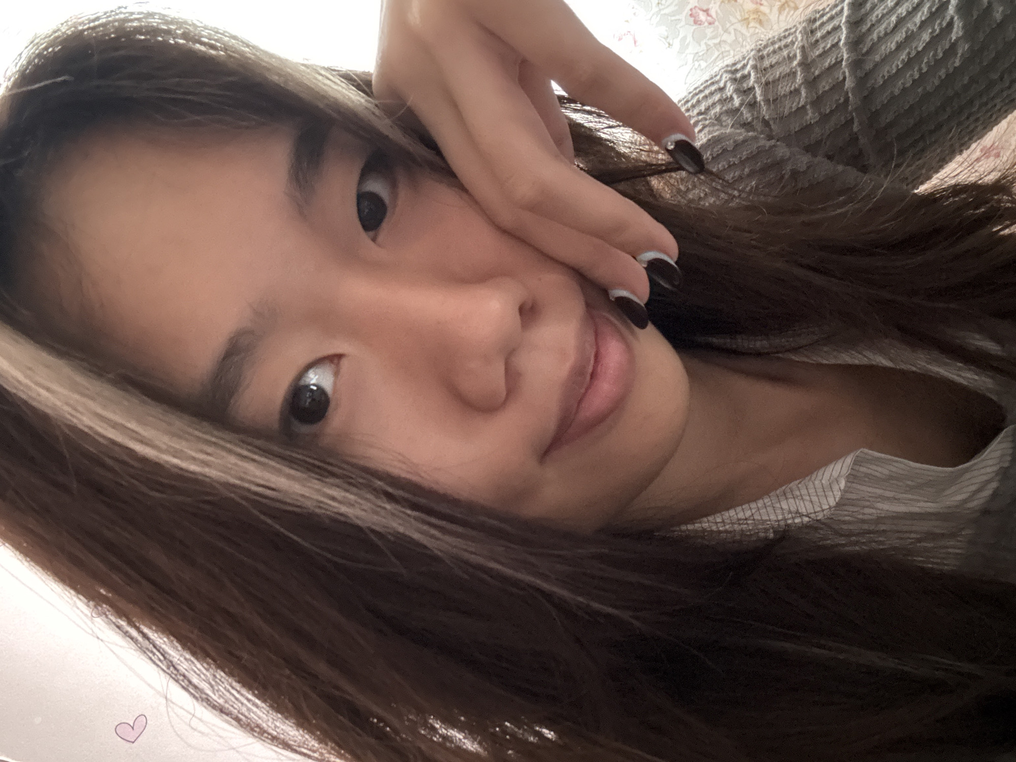 A young woman with long, dark brown hair is looking directly at the camera. Her left hand is partially covering her face. She has dark eyes, fair skin, and her nails are painted black with a thin white stripe at the base. She is wearing a gray sweater over a striped shirt. A small, faint purple heart graphic is in the lower-left corner. There is no contextual information to support a location in the city of Shapingba District, China.