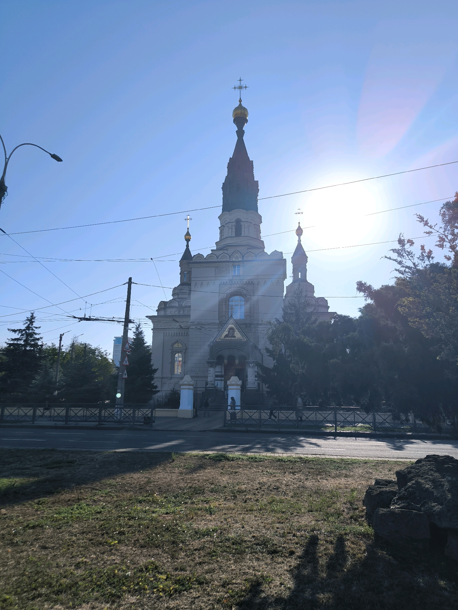 The image depicts the Church of St. Nicholas in Mykolaiv, Ukraine, on a clear, sunny day. The church, a prominent architectural landmark, features a tall, dark spire topped with a golden dome and cross, along with a lighter-colored main structure and smaller domes. The sun is bright, positioned high in the sky and slightly to the right of the church's spire, creating a lens flare effect and casting long shadows across the foreground.
In front of the church, there is a paved area with a dark metal fence. A street with a visible lane marking runs in front of the fenced area, and beyond that, a grassy slope with some dry grass and sparse green patches leads to the foreground. The slope is partially covered in shadows. There are a few silhouetted figures on the paved area in front of the church entrance, suggesting people are present.
To the left of the church, a street lamp with a curved arm is visible against the bright blue sky. Overhead, multiple power lines and utility wires crisscross the sky, partially obscuring the view of the church and the sky. Several evergreen trees are situated behind the fence and to the sides of the church, with some branches of a deciduous tree with reddish leaves visible on the right side of the frame. In the far background, there appears to be a taller, modern building visible through the trees. The overall scene is a street-level view of the church under a bright sky with urban infrastructure elements like power lines and a street lamp.