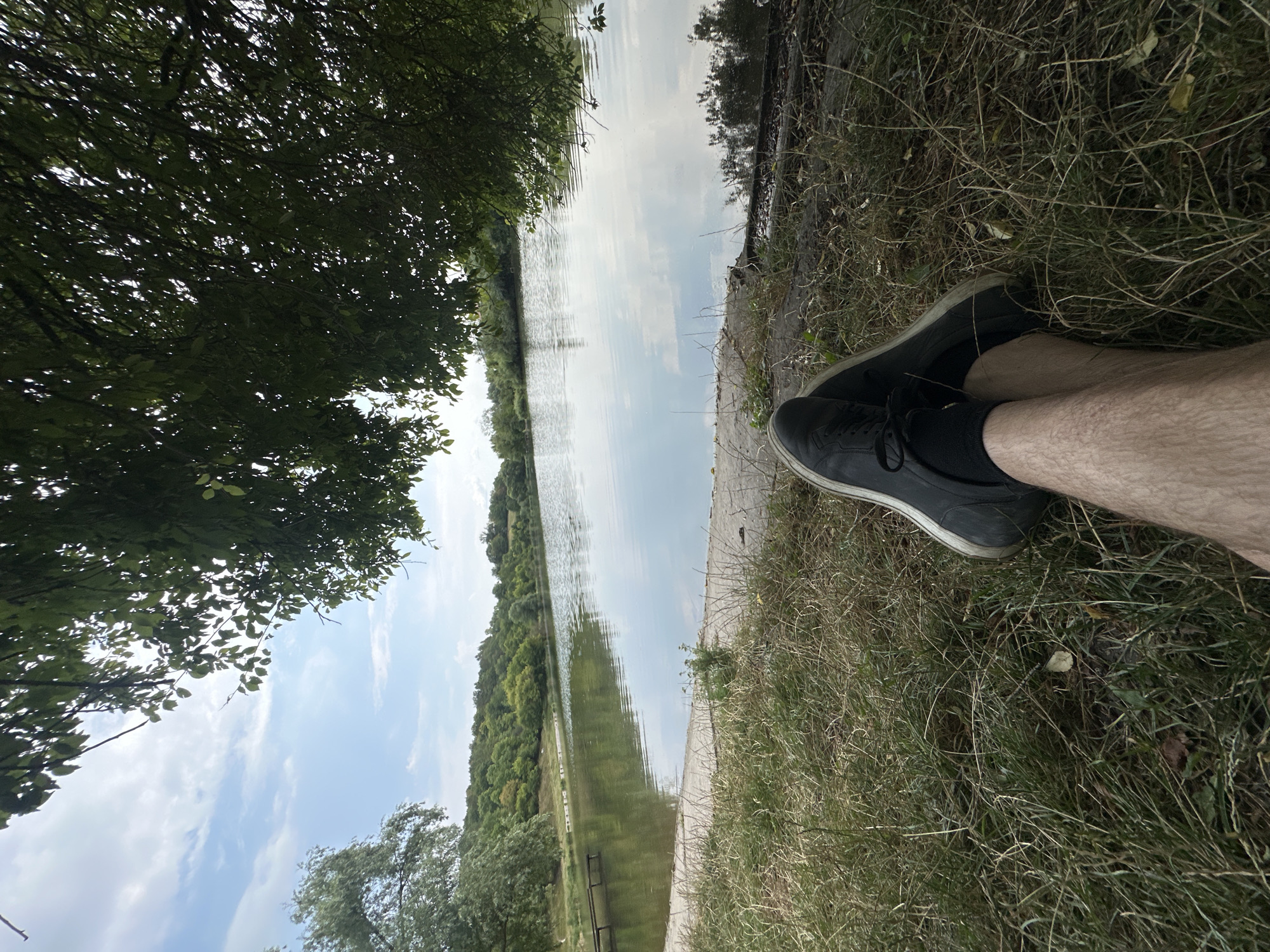 A low-angle outdoor shot shows a person's legs and feet resting in the foreground. The individual is wearing dark sneakers and black socks. They are seated on dry grass, which is illuminated by the sun. Behind the person is a body of water, possibly a lake or river, with its surface reflecting the sky and surrounding trees. The water is calm, with gentle ripples. On the opposite bank of the water, a lush green forest covers the rolling hills. The sky above is blue with scattered white clouds. The scene suggests a peaceful and natural setting, likely in the rural outskirts of Makiv, Ukraine.