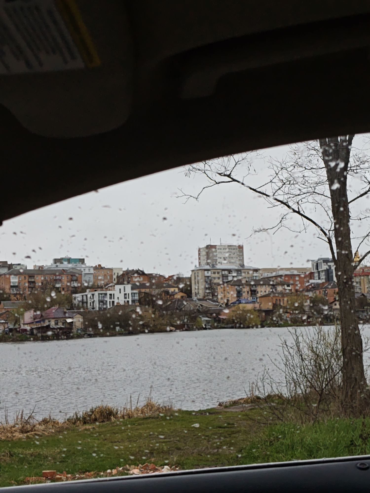 A view from inside a vehicle shows a cityscape across a body of water on a rainy day. The buildings are a mix of low-rise residential and taller apartment blocks, showing a variety of architectural styles and ages. A leafless tree stands to the right. The shore is grassy with some weeds and scattered bricks. Raindrops are visible on the windshield. The location is possibly in the city of Vinnytsia, Ukraine.