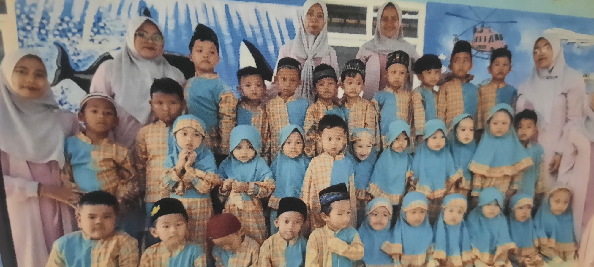 This is a full-shot photograph of a group of children and two adults, likely teachers, posing for a photo in what appears to be a classroom or school setting in Ploso, Indonesia. The background features a mural depicting a body of water with a whale's tail visible, and further to the right, a helicopter.
The group consists of approximately twenty-two children and two adults. The children are arranged in multiple rows, with the youngest appearing to be in the front. Most of the children are wearing matching outfits, consisting of a light blue top and pants with a tan and light blue plaid pattern, and a tan or light blue headscarf for the girls. Some of the boys are wearing peci hats in black or with decorative stitching. The two adults are in the back row, wearing light-colored hijabs and long garments, with one adult wearing glasses.
The children are looking towards the camera, and their expressions are varied, ranging from neutral to slightly smiling. Some of the younger children in the front row are looking around or slightly off-camera. The adults appear to be smiling.
The overall impression is of a formal school or kindergarten photo, likely taken during a special event or the beginning of a school year. The setting suggests a children's educational institution.
