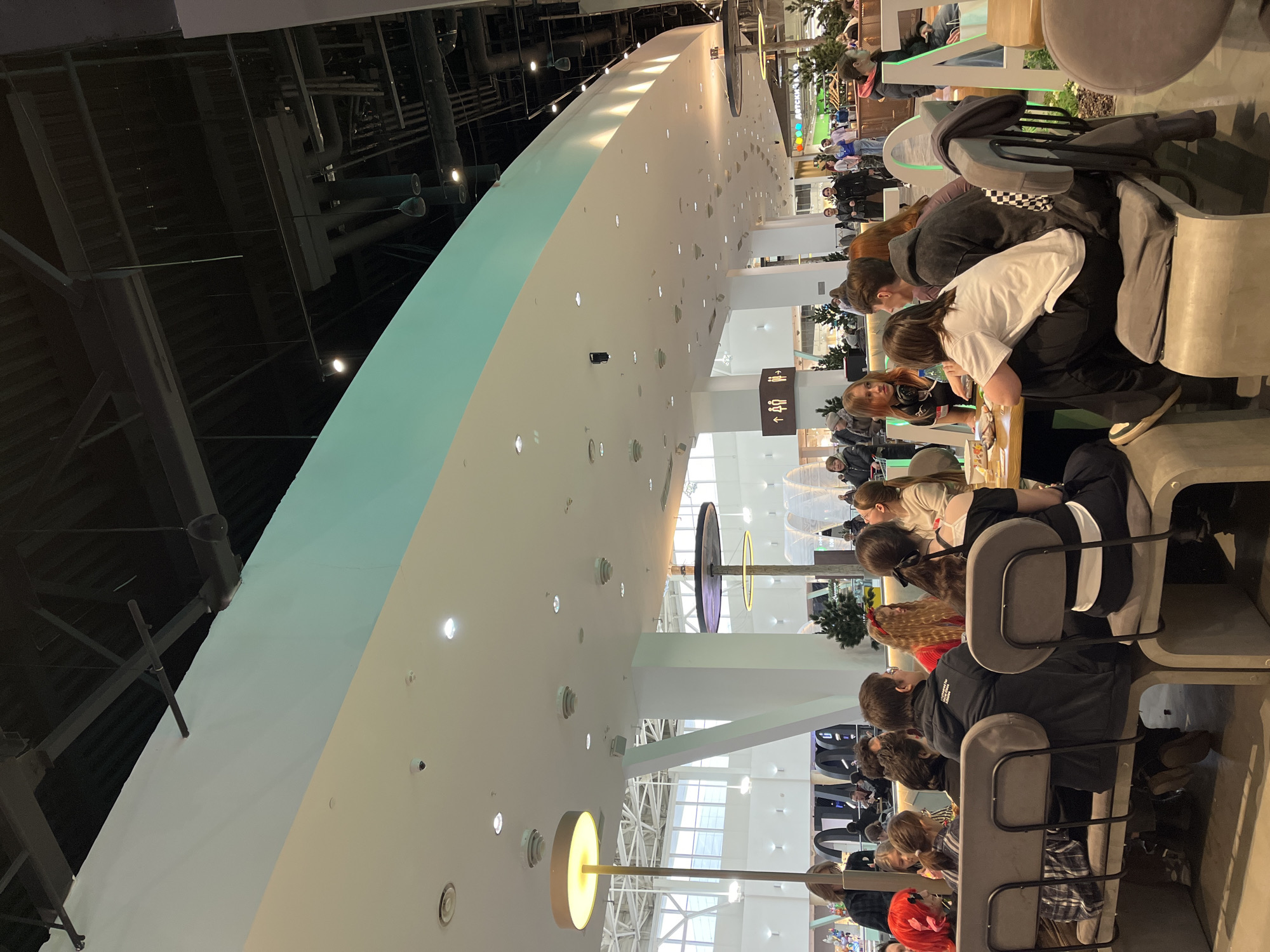 This image depicts a bustling indoor scene, likely a modern food court or cafe area within a large public building or shopping mall in Krasnoyarsk, Russia. The perspective is taken from a slightly elevated position, looking down a long, curved seating arrangement.
The primary subjects are numerous people, appearing to be predominantly young adults and teenagers, seated at long, light-colored tables. They are occupying grey, upholstered chairs and engaged in various activities such as eating, conversing, and possibly using electronic devices. Their attire is casual and contemporary. One person in the foreground with reddish-brown hair glances towards the camera.
The setting is characterized by a distinctive modern design. A prominent, smooth white curved wall with recessed lights dominates the left side of the frame, transitioning to a light blue-green section towards the top. Above this structure, the ceiling features an industrial aesthetic with exposed dark pipes, ventilation ducts, and structural beams. In the background, large windows allow natural light, and decorative circular light fixtures are suspended from the ceiling.
Notable details include a clear brown sign on a white pillar displaying universal restroom symbols (male, female, disabled access, baby changing) with an upward arrow. Partially visible Cyrillic text in the distant background near a colorful display likely reads "ДЕТСКИЙ" (Children's). The overall atmosphere appears casual and lively, suggesting daytime hours due to the bright artificial and visible natural light. No specific event is evident beyond general social interaction and dining in a public space.