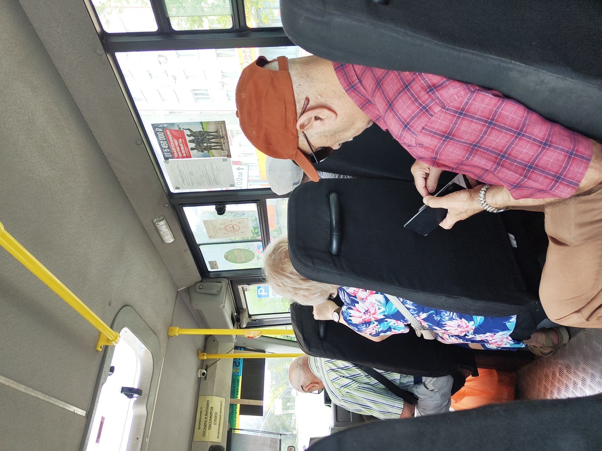 The image depicts the interior of a bus, likely in Ramenskoye, Russia, as suggested by context. In the foreground, a man wearing a red plaid shirt, brown baseball cap, and sunglasses is seated, looking down at his phone. He has a watch on his left wrist. Behind him, to his left, a woman with blonde hair wearing a floral dress is also seated. Further to the left, a man in a striped shirt and glasses is visible. The bus interior features black seats, yellow handrails, and a window displaying buildings and trees outside. Signs are visible on the interior walls and windows.