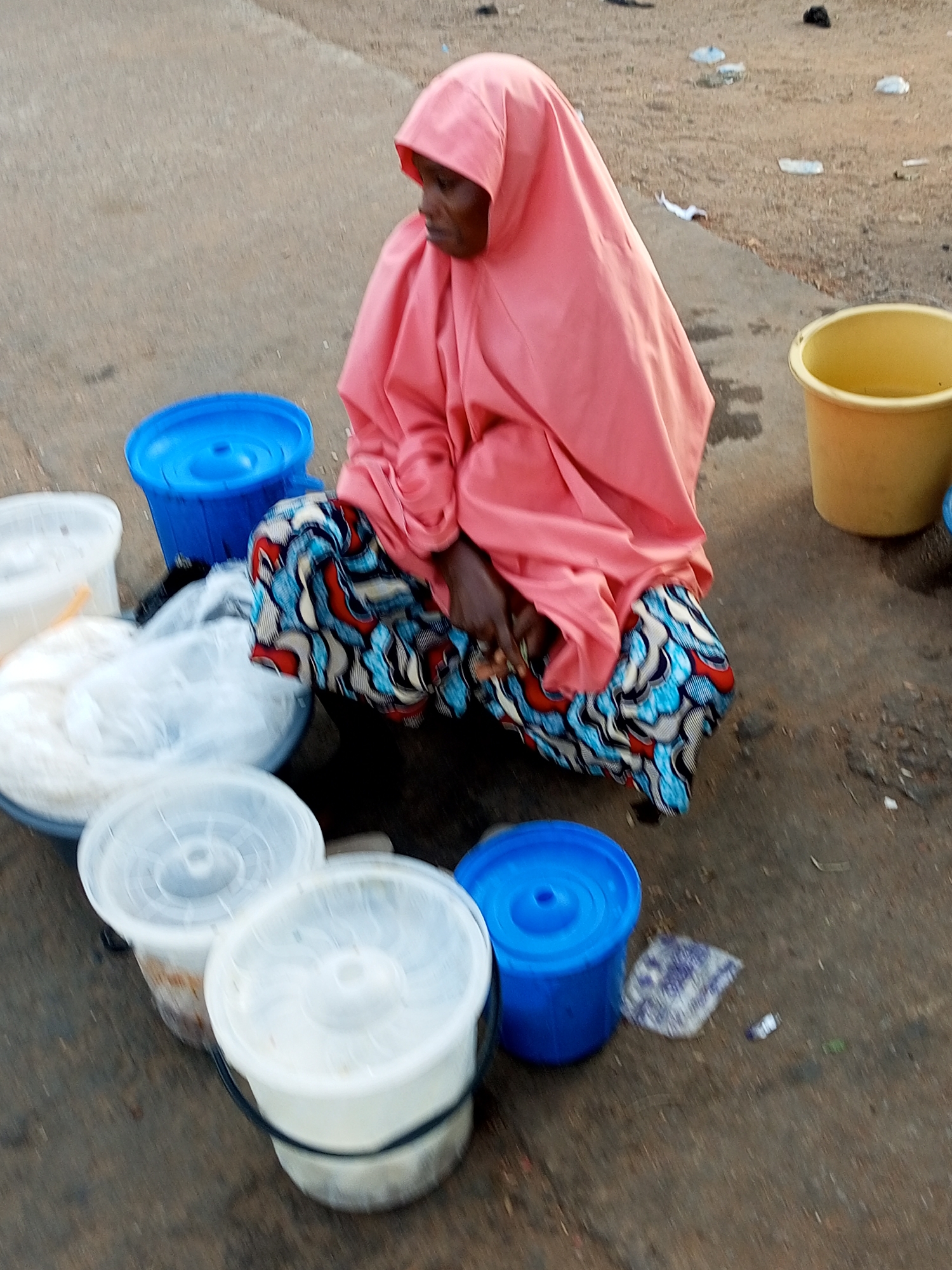 In Gombe, Nigeria, a person is crouching beside several large plastic containers filled with goods, likely for sale. The individual, dressed in a pink headscarf and a patterned skirt, is positioned on a rough, dusty ground surface that appears to be a street or market area. Surrounding the person are various containers, including a yellow bucket, blue buckets, and white plastic tubs with lids. The overall scene suggests a commercial activity taking place outdoors in a public space.