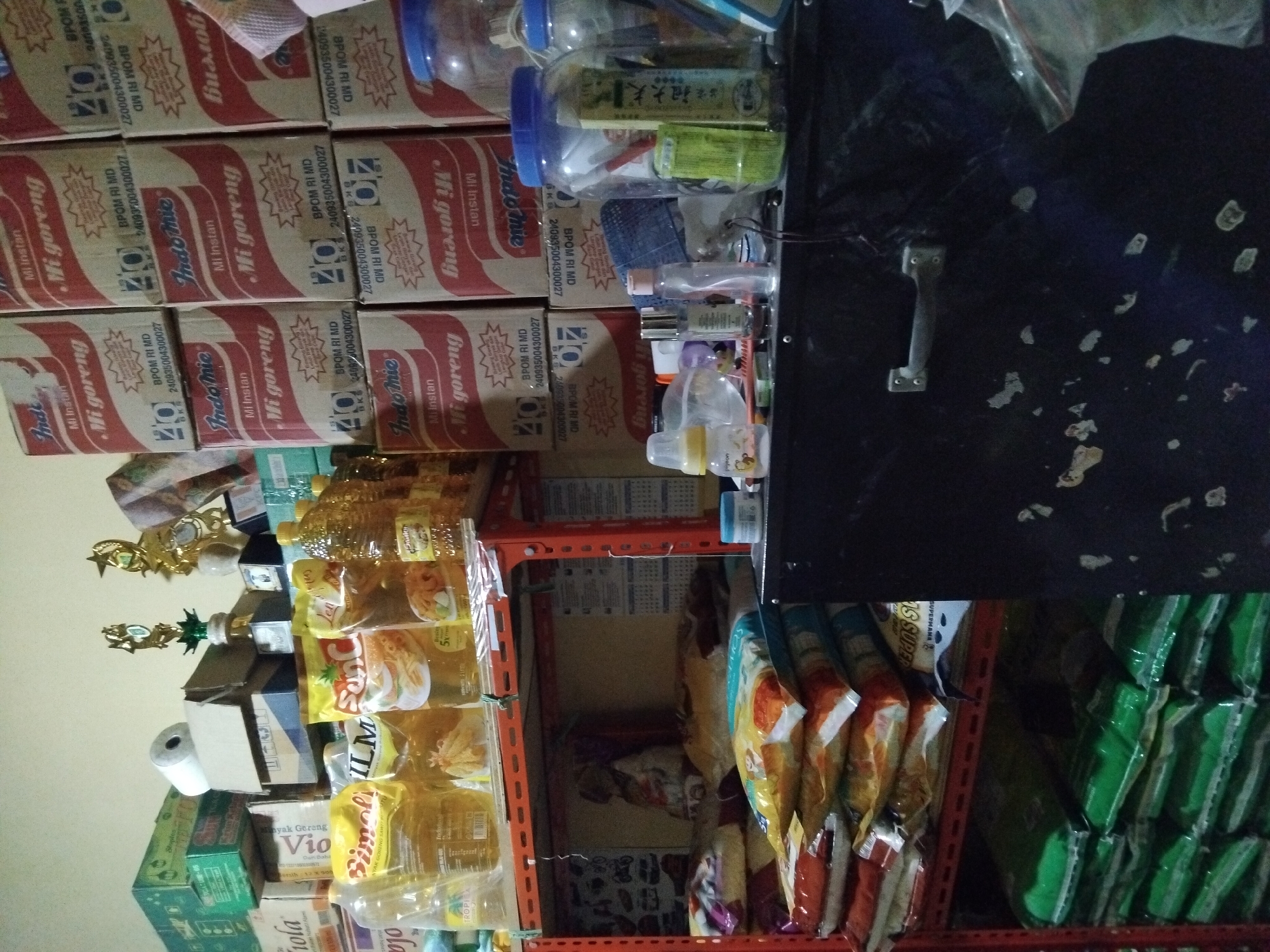 The image shows a shelf filled with various food items and household supplies in what appears to be a storage area or a small shop in Kepuh Kiriman, Indonesia. On the top shelf, there are several large plastic bottles of cooking oil, packages of instant noodles (Indomie Mi Goreng), and other food products. There are also some decorative trophies on a shelf above the food items. The middle shelf contains more food items, including bags of rice and smaller packages of food. On the bottom shelf, there are also stacked bags of rice. To the right of the shelves, there is a black cabinet with a handle. The overall scene suggests a retail or storage environment where various consumer goods are kept.