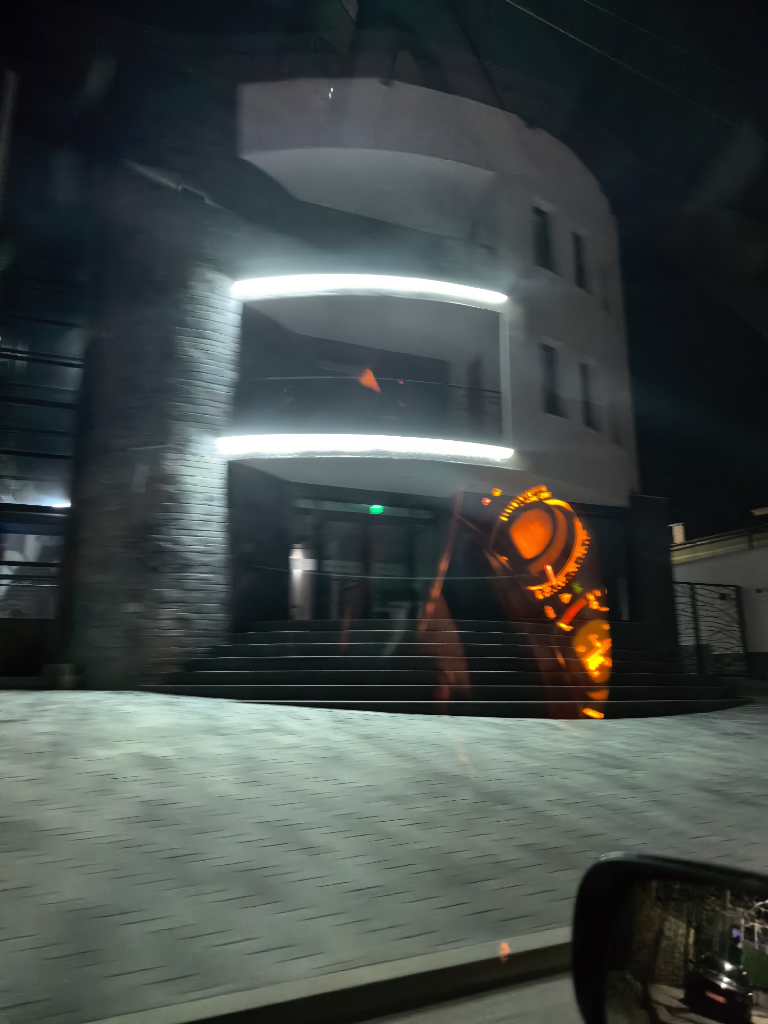 The image, taken at night, depicts a building with a modern design. The building has a façade that combines brickwork and smooth white surfaces. It is illuminated by horizontal light fixtures that cast a bright glow. There is a series of steps leading to the entrance of the building. An orange and red illuminated design is visible next to the building's entrance. The photograph appears to have been taken from inside a vehicle, as part of the car's side mirror is visible in the lower right corner. The street in front of the building is paved with light-colored bricks.