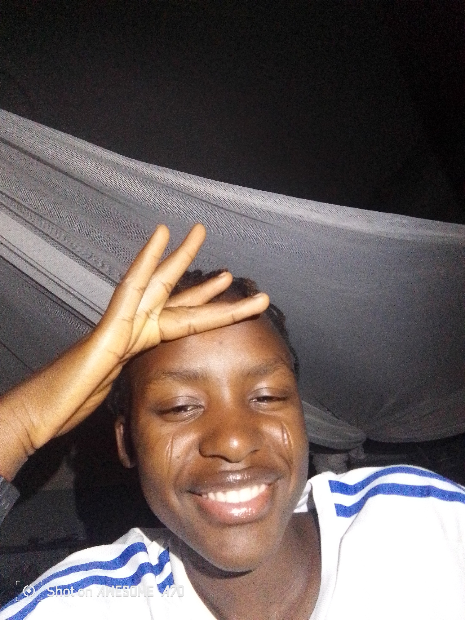 A close-up shot shows a person smiling with their hand on their forehead. They are wearing a white shirt with blue stripes on the shoulder. A mosquito net is visible in the background. The location is Unknown, Unknown.