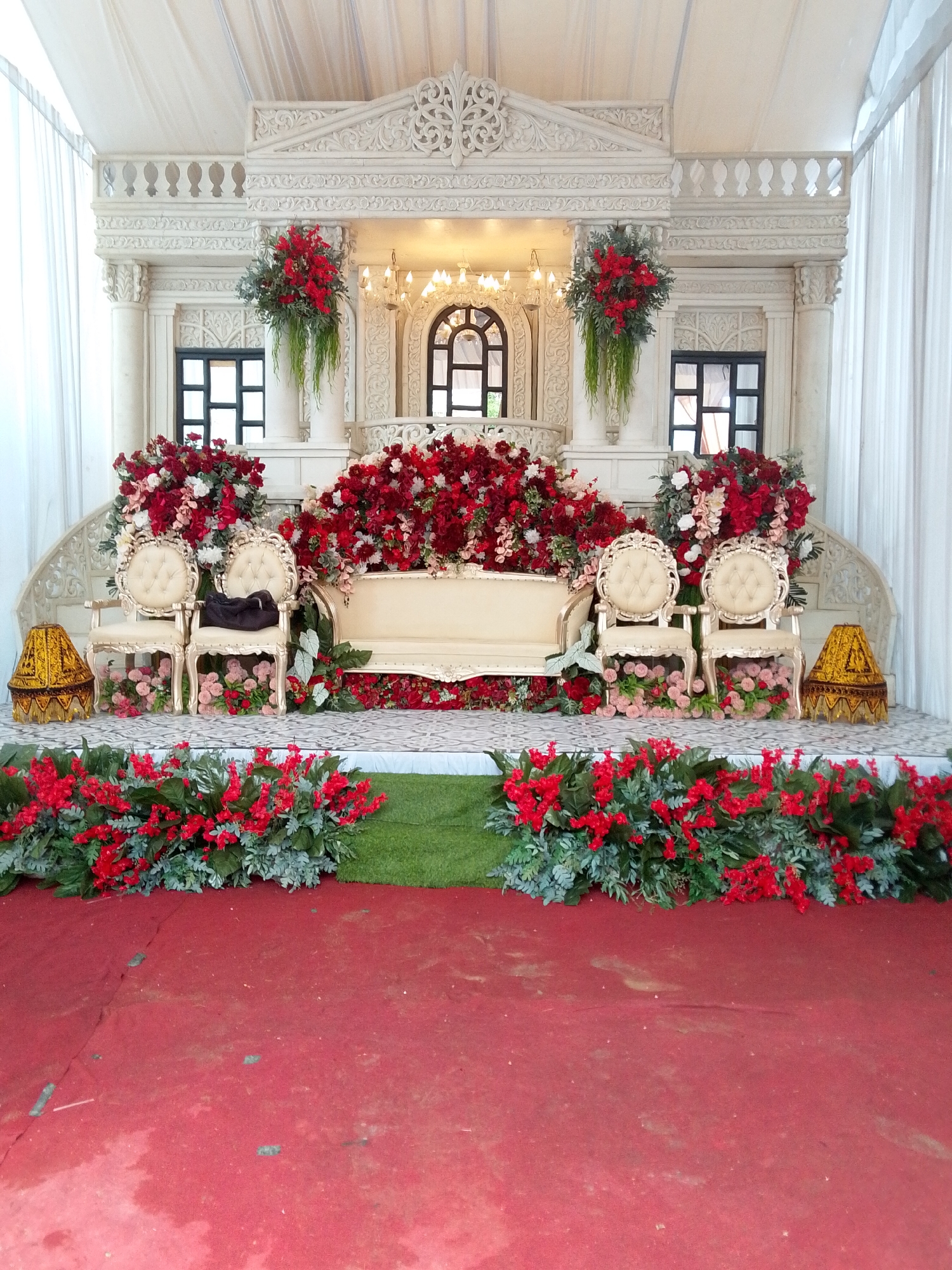 The image shows a wedding stage in Payakumbuh, Indonesia. The stage is adorned with white ornate structures resembling classical architecture, draped with white fabric. A large arrangement of red and white flowers dominates the center, with two ornate cream-colored chairs positioned in front of a cream-colored sofa. A decorative gold lamp is placed to the left of the chairs, and another similar lamp is on the right, partially obscured by flowers. Lush greenery and red flowers are arranged around the base of the stage and extend onto the red carpeted floor. The overall scene suggests a formal and elaborate wedding ceremony setting.