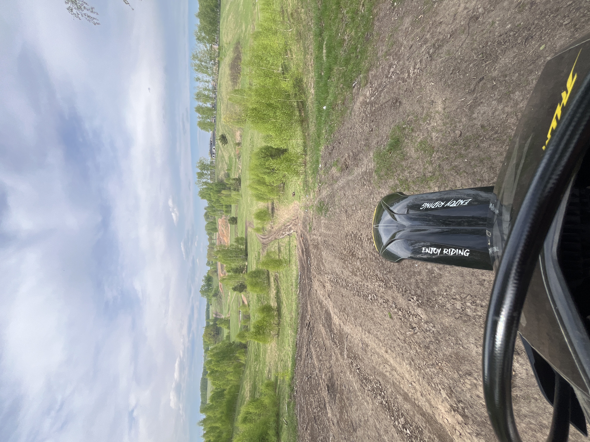 This image is a first-person view from a vehicle, possibly an ATV, moving along a dirt track. The vehicle's dashboard, with "ENJOY RIDING" written on it, and part of the handlebars are visible in the foreground. The dirt track extends into the distance. To the left of the track is a grassy area with scattered trees. Further in the distance, a line of trees is visible, possibly indicating the edge of the area. The sky, featuring a mix of blue and white clouds, is visible in the background. Based on other sources, the location is the city of Новочувашский, Russia.