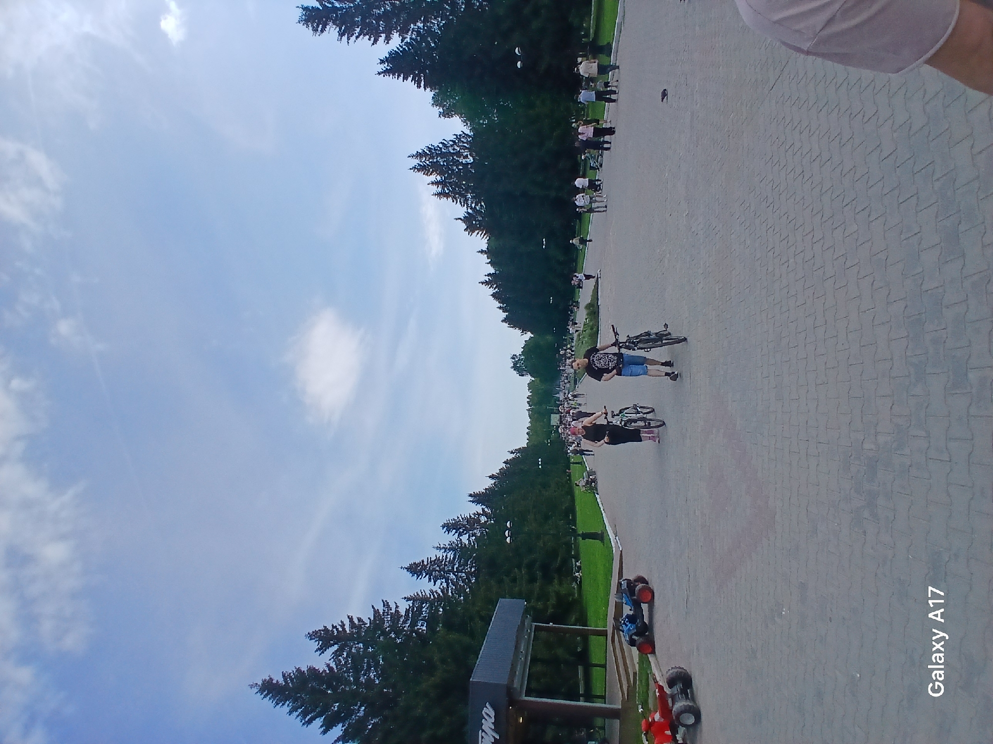 A public park scene in Yekaterinburg, Russia shows a wide paved walkway lined with mature trees on the left and various passersby. Two people are walking bicycles. There are at least three small toy cars or ride-on toys on the walkway. People are strolling in the distance. The sky is blue with some clouds.