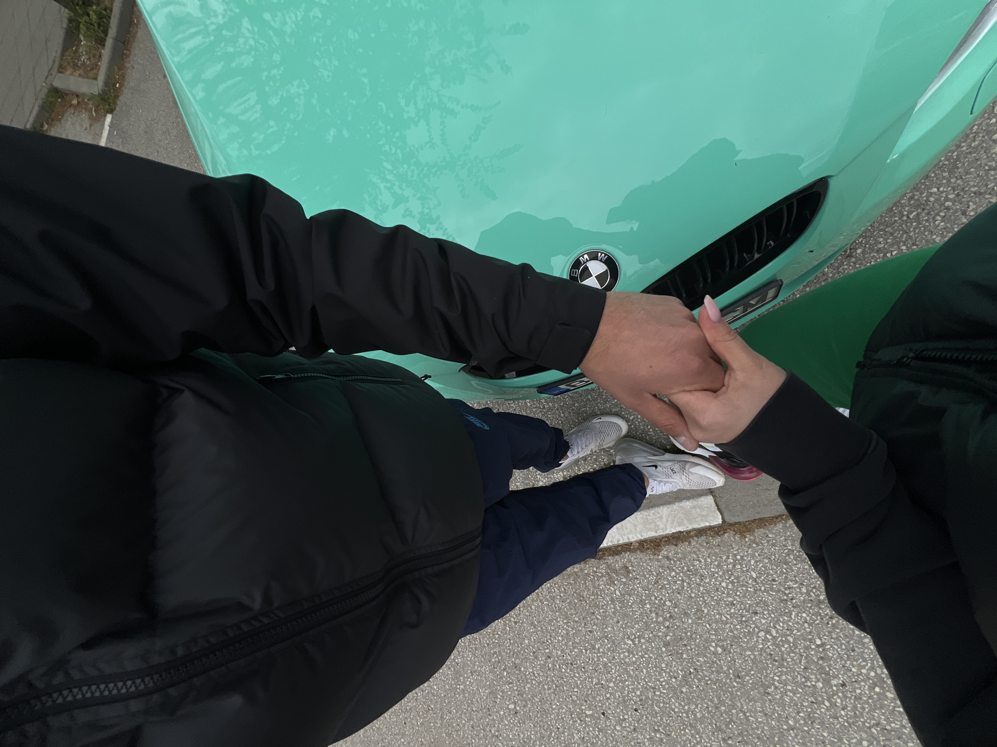 An overhead shot captures two individuals holding hands, standing next to a vibrant turquoise BMW car. The scene appears to be outdoors on a paved surface, possibly in Plovdiv, Bulgaria.
The person on the left wears a black puffer jacket over a black long-sleeved top, dark blue pants, and white Nike Air Max 270 athletic shoes with gray details. Their right hand is interlocked with the left hand of the person on the right. This second individual wears a black long-sleeved top, a dark green lower garment, and white Nike Air Max 270 sneakers featuring pink accents. Light pink nail polish is visible on their fingers.
The striking turquoise car occupies the upper portion of the frame. Its hood clearly displays the circular BMW emblem, and a black automotive grille or vent, possibly from an "M" series model, is visible on the right side of the hood. Reflections, potentially from overhead trees or a cloudy sky, are apparent on the car's surface. The ground is a light gray, pebbled asphalt or concrete, with a concrete curb visible beneath the feet of the person on the left. The visible text includes "BMW" on the car's emblem and "Nike" and "270" on the shoes. The overall impression is a daytime scene, possibly capturing a casual moment of togetherness.