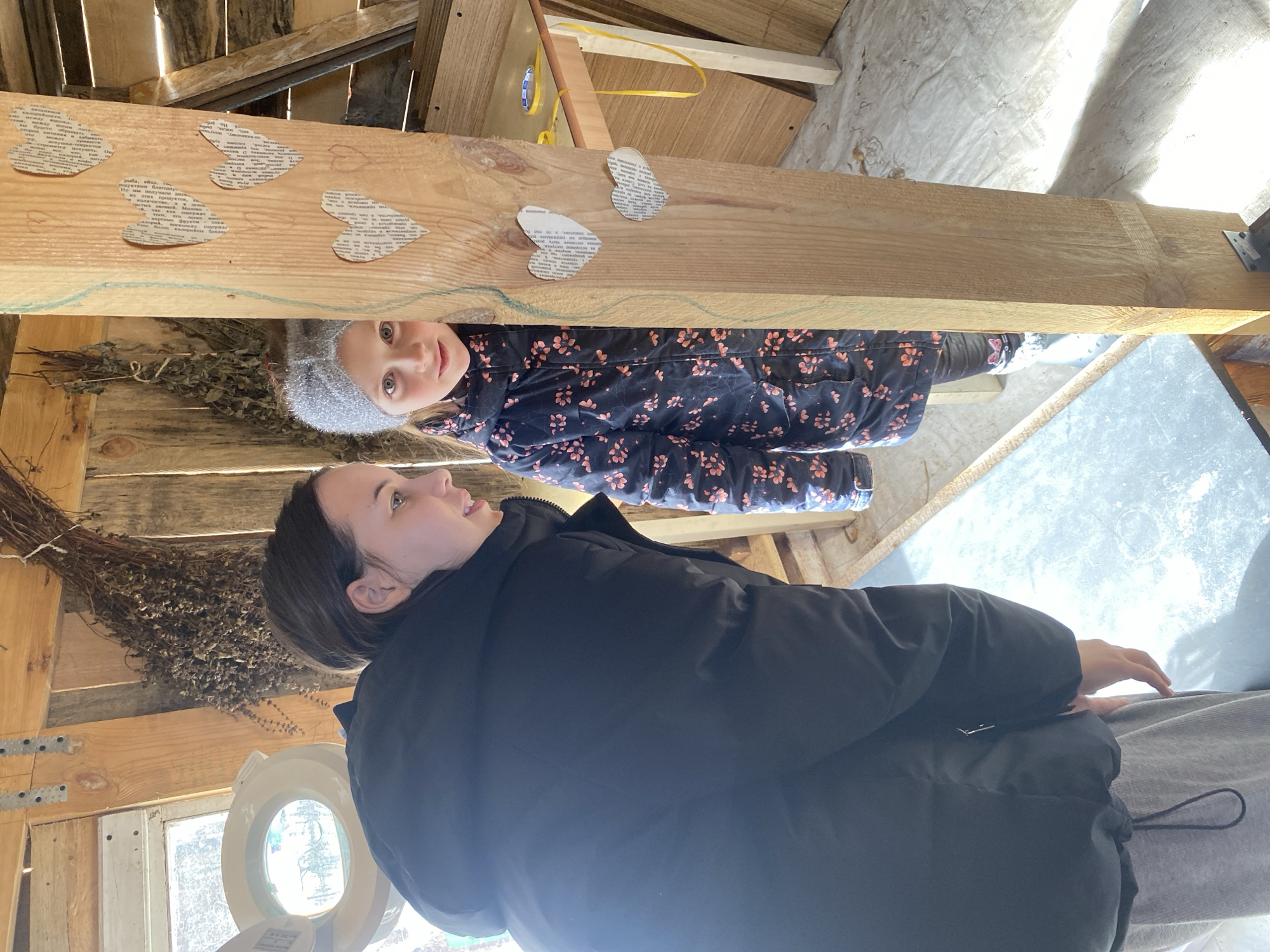 This image captures an interior scene within a rustic wooden structure in Noviy, Russia. An adult female, positioned on the left, is seen from the torso up, wearing a black puffer jacket and light grey sweatpants, leaning forward and looking to the right. To her right, a young girl is partially visible, peeking from behind a prominent vertical wooden post. The girl wears a grey knitted beanie and a dark winter jacket with a pink floral pattern, looking directly at the camera with a slight smile. The wooden post is adorned with multiple heart-shaped cutouts made from text-printed paper, along with faint drawn heart outlines. The background features walls constructed from rough wooden planks, with bundles of dried plants hanging from the upper section. A window in the upper left allows bright sunlight into the space, alongside a circular white fixture. To the right of the wooden post, a light-colored wooden table or surface holds a roll of blue adhesive tape and a yellow ribbon. The floor, illuminated by sunlight, appears to be a light-colored, possibly concrete or covered, surface.