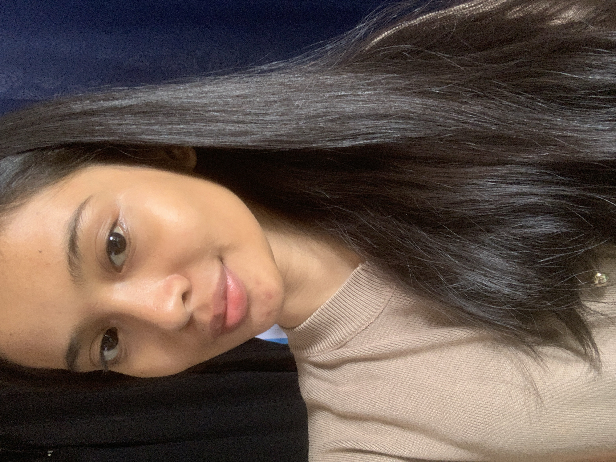 The media file is a portrait of a young woman with dark hair. She is leaning forward, with her face turned towards the camera. Her eyes are open and looking at the viewer, and she has a slight smile on her lips. Her hair is long and dark, and it falls around her shoulders and down her back. She is wearing a beige, ribbed sweater. The background is dark and out of focus, with a hint of blue in the upper left corner. There are no visible objects or text in the image. The lighting suggests an indoor setting, possibly at dusk or with artificial light. The woman appears relaxed and confident.