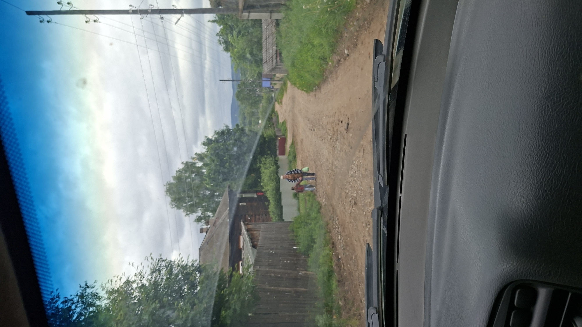 The image shows a dirt road in a rural village, likely in Novoilinsky, Russia. The road is lined with wooden houses and fences, with greenery on either side. Two people, an adult and a child, are walking down the road away from the camera, holding hands. The adult is carrying a green plastic bag. The sky is cloudy. The foreground shows the dashboard and windshield wipers of a car.