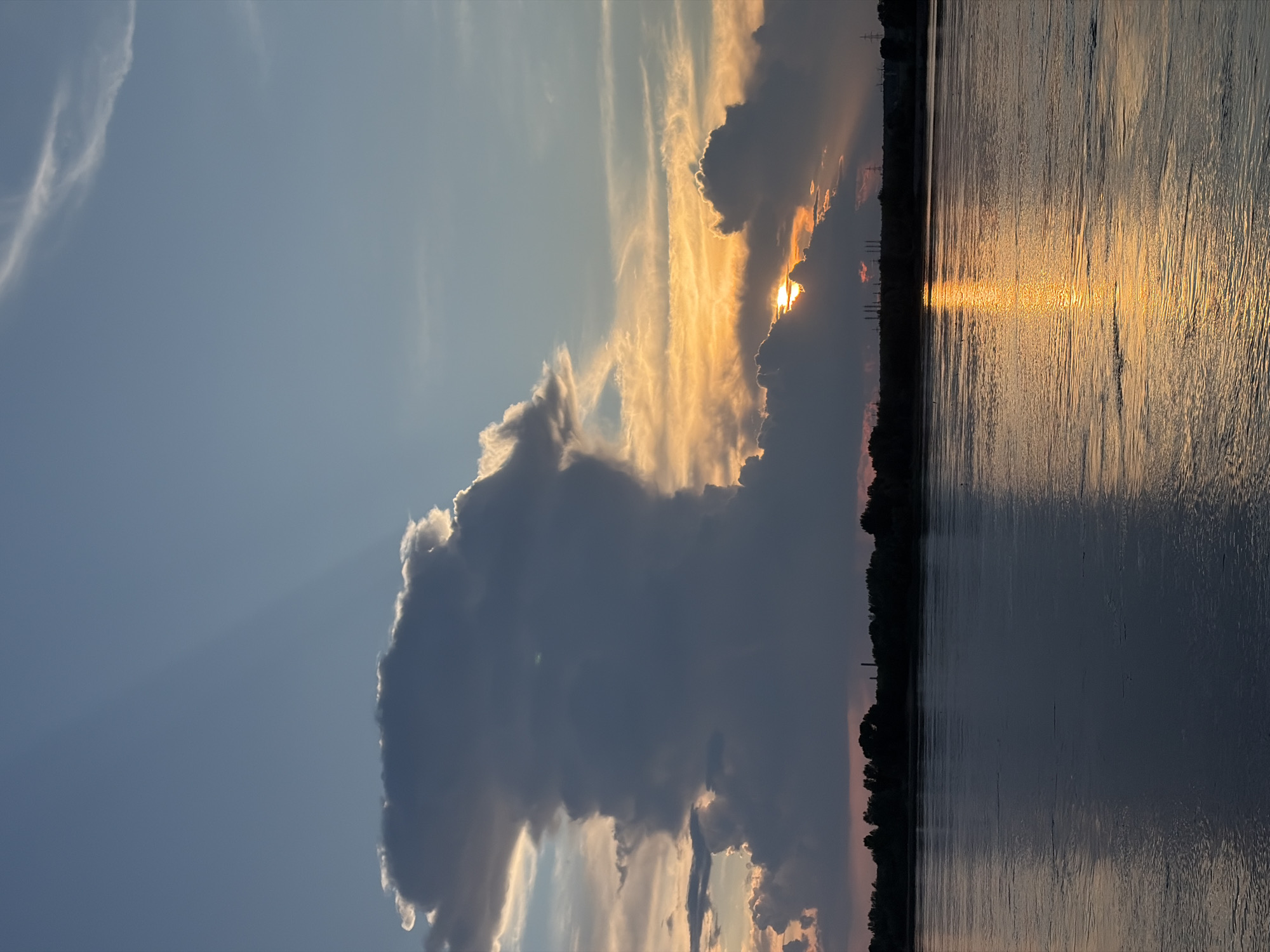 A vertical, low-angle shot captures a dramatic sunset over the Kama River in Chaikovsky, Russia. The sky transitions from a clear, pale blue at the top to a vibrant orange and yellow near the horizon, where a large, dark cumulus cloud dominates the scene. Rays of sunlight pierce through gaps in the cloud cover, casting light and shadow. Below the sky, a dark silhouette of trees lines the riverbank, interrupted by industrial structures. The river itself reflects the fiery hues of the sunset, with ripples and currents visible on its surface. The overall mood is serene yet powerful, marked by the contrast between the dark, imposing cloud and the bright, warm sunset.