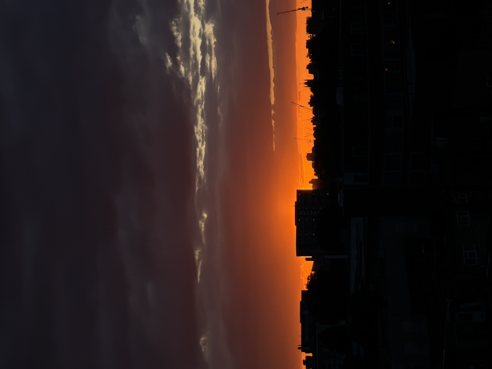 The image depicts a dusk or dawn scene in what appears to be a city, silhouetted against a dramatic sky. The silhouettes are likely buildings, with possible construction cranes visible on the horizon. The sky transitions from a dark area on the left, possibly indicative of heavy cloud cover, to a bright orange and yellow glow near the skyline, suggesting either sunrise or sunset. The location might be identified based on the skyline's distinctive features, although such identification would require expertise on London architecture and city planning. No people or specific interactions are visible in the image. The general environment is an urban landscape undergoing construction.