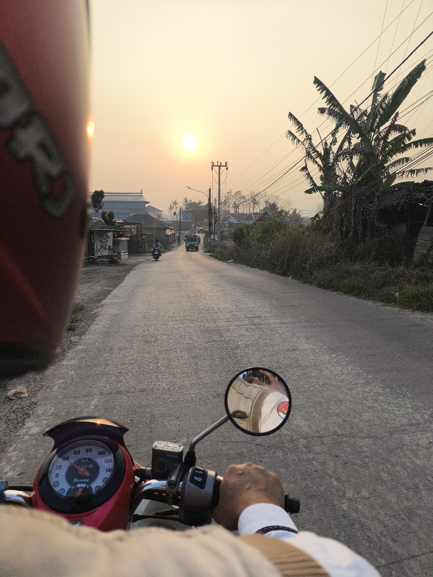 This image captures a first-person perspective of someone riding a red scooter on a paved road at sunset in Cilegon, Indonesia. The sun is low on the horizon, casting a warm, orange glow across the sky and illuminating the road. In the foreground, the rider's hand is visible on the handlebars, and a red scooter dashboard with a speedometer is in focus. A helmet, partially visible on the left, is also red.
The road ahead is lined with vegetation, some buildings, and utility poles with visible power lines. On the right side of the road, there are banana trees and a rustic structure with a thatched roof. Further down the road, a green truck and another scooter carrying two people can be seen. The rider's reflection is visible in the scooter's side mirror, showing them wearing a light-colored garment. The overall scene suggests a typical commute or journey through a semi-urban or rural area of Cilegon during the golden hour.
