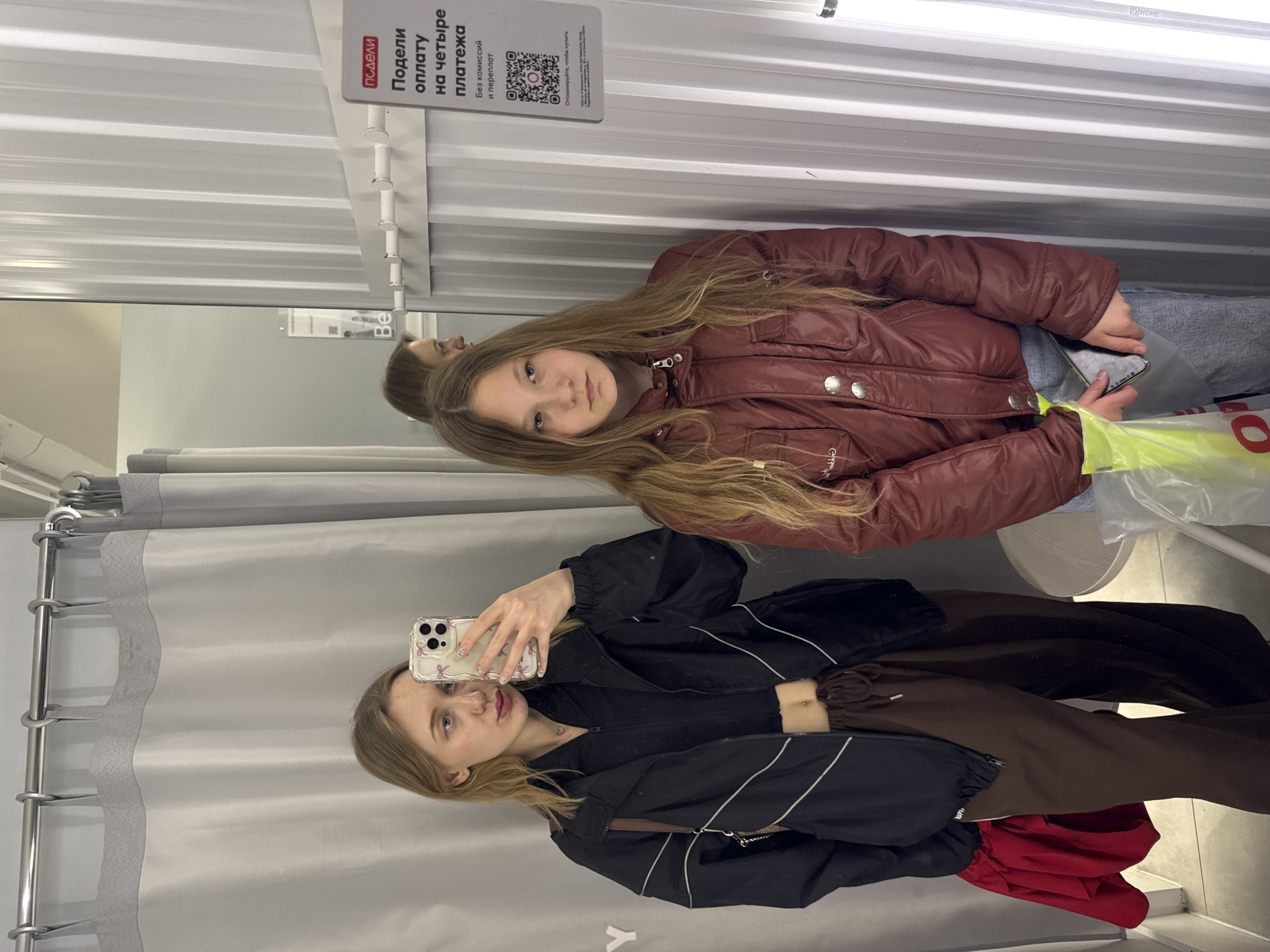 The image captures a young woman taking a selfie in a fitting room. The reflection in the mirror shows a second person, possibly a younger girl, posing with a burgundy jacket. The fitting room has gray curtains and a sign with the text "Подели оплату на четыре платежа" and a QR code. The environment appears to be a clothing store. The location is speculated to be Ovoshchnoy, Russia. The woman in the foreground is wearing a black jacket with white stripes, brown pants, and holding a phone with a patterned case. The girl in the reflection is wearing the burgundy jacket and blue jeans. There's a yellow and white bag visible on a chair.