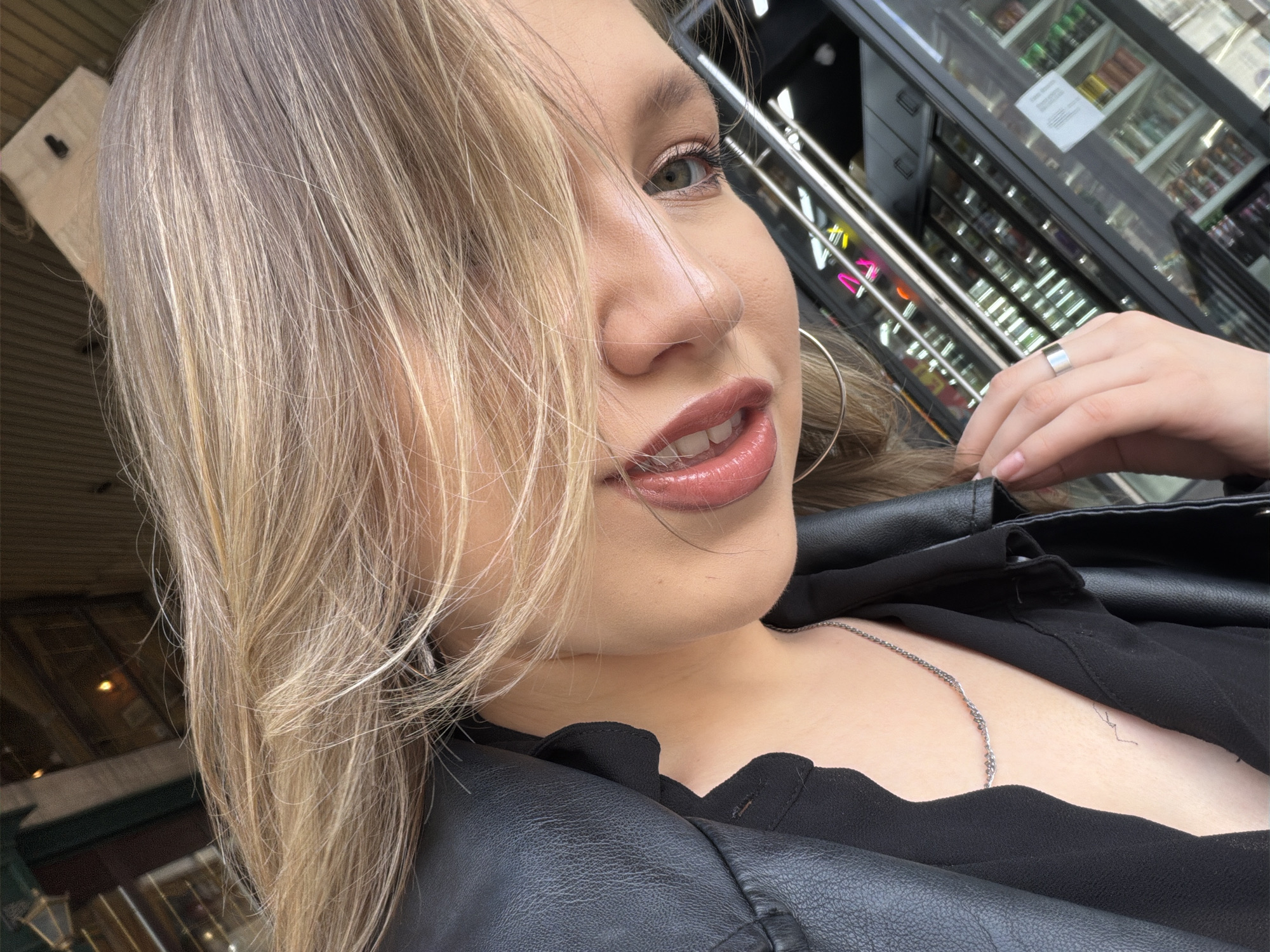 A close-up selfie shows a young woman with blonde hair, which partially covers her face. She has light green eyes and is wearing makeup, including lipstick with a glossy finish. She is also wearing a black leather jacket over a black top, and a silver chain necklace is visible. A silver ring is on her right hand. In the background, a glass-fronted display case with shelves of products is visible, along with some metallic structures and neon lights. The overall lighting suggests it is daytime. The scene appears to be in a public place with commercial activity.