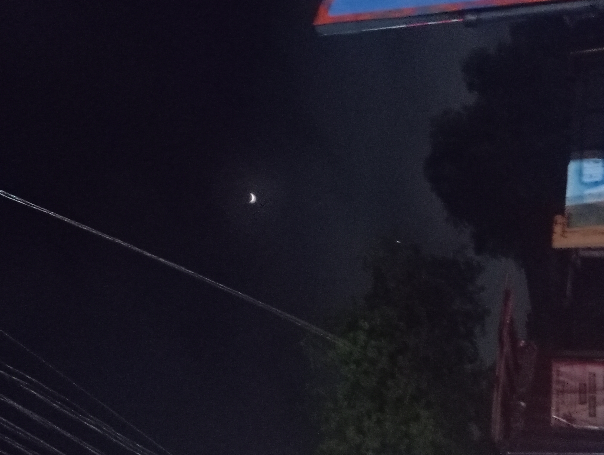 The image shows a night scene in what appears to be an urban environment, possibly Nkpor, Nigeria. A crescent moon is visible in the dark sky, slightly left of center. Power lines run across the left side of the frame. To the right, trees and the corner of a building with signage are visible, though details are obscured by the darkness. The overall lighting is poor, creating a low-visibility environment.