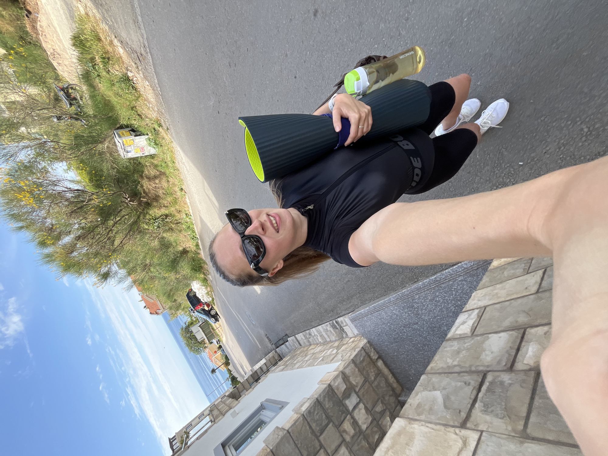 A woman is taking a selfie while holding a yoga mat and a water bottle on a street in Bečići, Montenegro. She is wearing a black athletic top and black shorts, along with white sneakers and sunglasses. The background includes vegetation, houses, the sea, and the sky. A street and a brick structure are also visible.