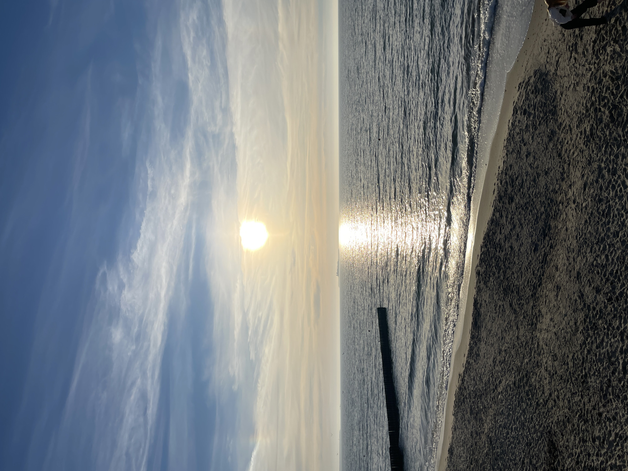 The image depicts a beach scene, likely at sunset, in Zelenogradsk, Russia. The view is oriented vertically. On the left, the sky is blue with wispy white clouds. The sun is positioned near the horizon, emitting a bright glow. The sea occupies the right half of the image, with a path of light reflecting off the water's surface. Wooden breakwaters extend into the sea. The shoreline consists of sand, and waves are visible near the edge. A person is visible in the top right corner.