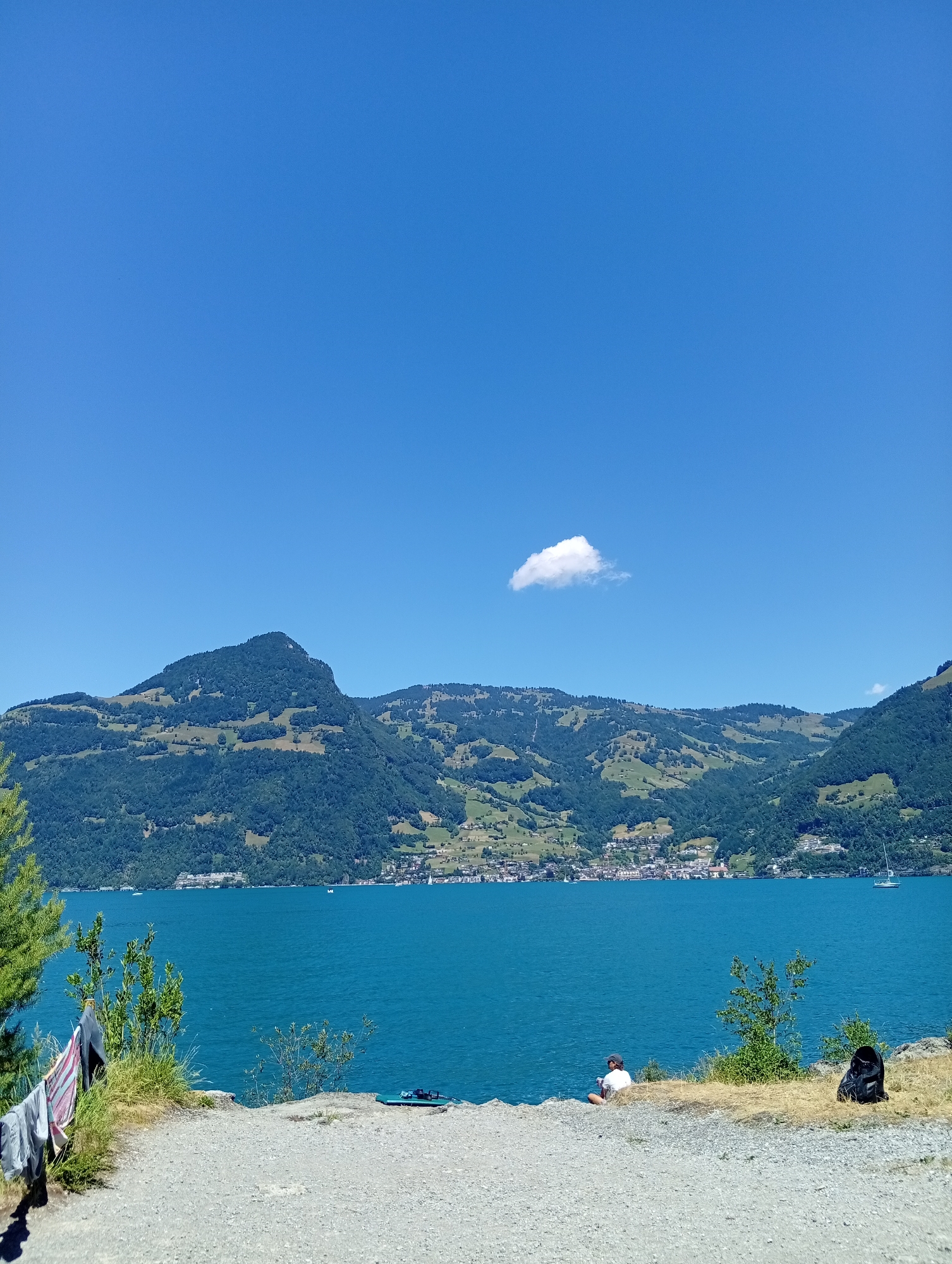 The image depicts a serene lake scene with a person sitting on the shore in Emmetten, Switzerland. The sky is a clear, vibrant blue with a single, small white cloud. Dominating the background are lush green mountains dotted with buildings and clearings, their slopes intricately terraced. The turquoise water of the lake stretches across the mid-ground, reflecting the bright sky. In the foreground, a gravelly path leads down to the water's edge, where a person in a white shirt and dark shorts is seated on the grass, looking out at the lake. To their right, a black backpack rests on the dry grass. To the left of the path, laundry hangs on a makeshift line, adding a touch of domesticity to the otherwise natural landscape. Boats are visible scattered across the lake, suggesting recreational activity. The overall impression is one of a peaceful, sun-drenched day in a picturesque Swiss setting.