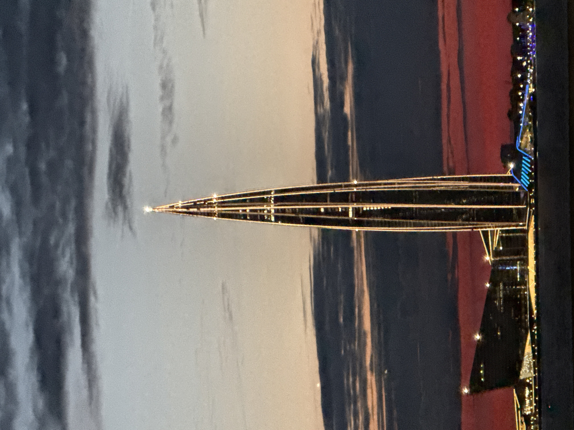 The image shows the Lakhta Center, a skyscraper located in the northwest of Saint Petersburg, Russia, on the Gulf of Finland. The photograph captures the tower at dusk, with the sky displaying a gradient of colors from pale grey at the top to a vibrant red near the horizon. Dark, wispy clouds are scattered across the sky. The Lakhta Center, illuminated by lights that trace its spire-like structure, dominates the center of the frame. In the foreground, a body of water, likely the Gulf of Finland, is visible. On the left and right sides of the image, parts of the city's waterfront are illuminated, including a distinctively shaped building with a dark facade and glowing windows, and a structure with bright blue accents. The overall scene depicts an urban landscape at twilight with a focus on architectural illumination against a dramatic sky.