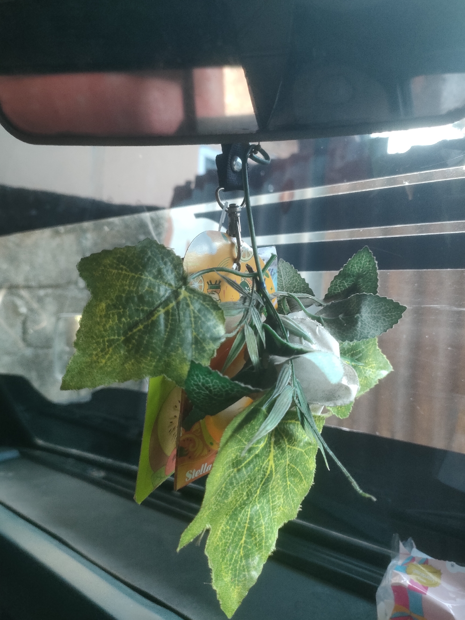 The image shows a close-up view of a car's rearview mirror with a decorative hanging item attached to it. The hanging item consists of artificial green leaves and a white flower. Behind the mirror, the reflection of a person's arm and shoulder is visible, suggesting they are the driver or a passenger. The background appears to be a window with horizontal blinds. The scene is indoors, inside a vehicle. There is no information to indicate the location in the city of Padang Dalom, Indonesia.