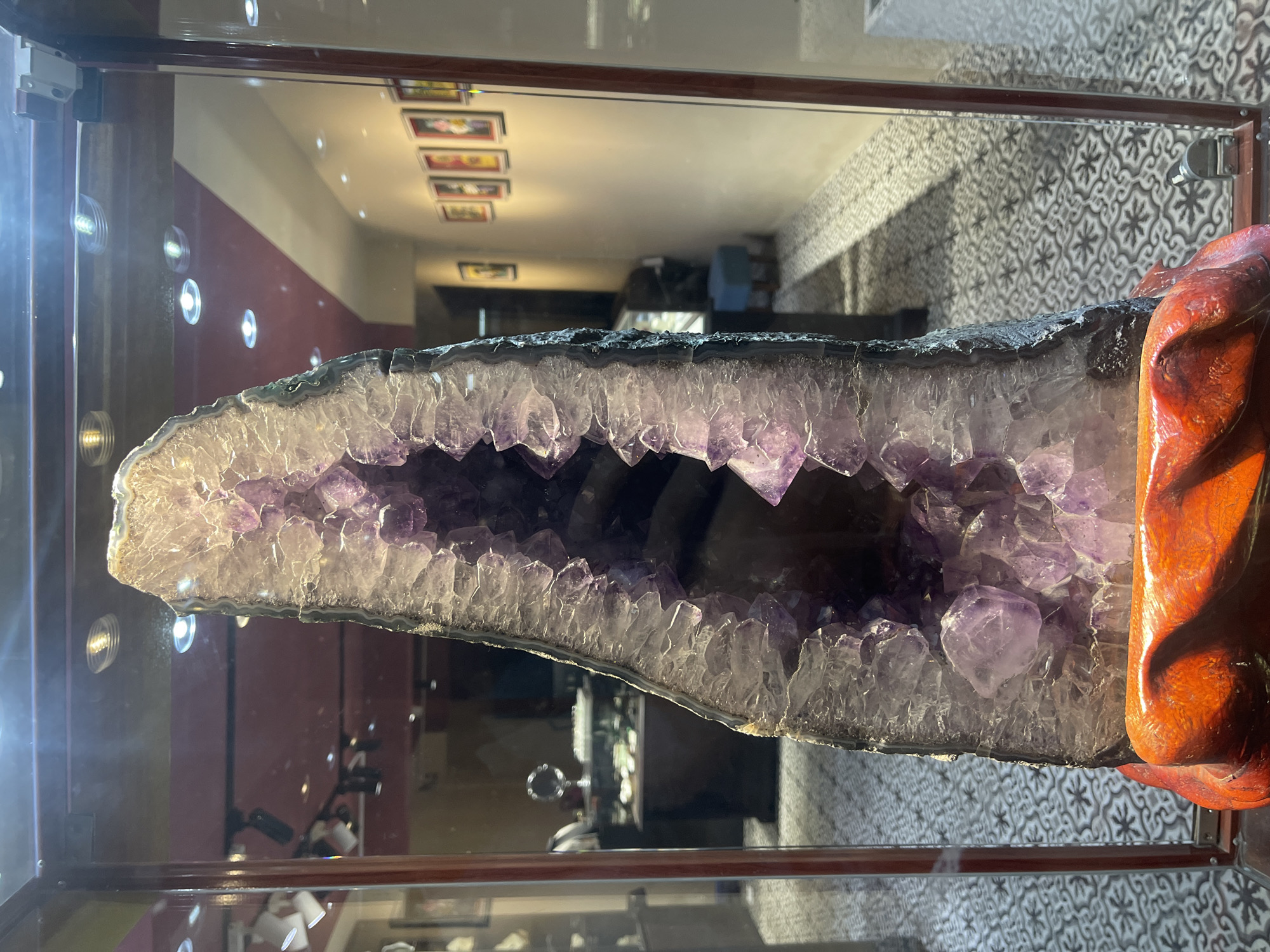 The image presents a large amethyst geode displayed within a reflective glass case or window, likely in a shop or gallery setting in Nha Trang, Vietnam. The geode is the central subject, a substantial rock formation split open to reveal an interior cavity densely lined with numerous purple amethyst crystals. These crystals vary in size and intensity of color, ranging from lighter translucent hues to deeper, richer purples. The outer shell of the geode appears dark and rugged.
To the right, the geode is supported by a distinctive, intricately carved wooden stand, which is reddish-brown and glossy. The reflections on the glass surface are prominent, revealing details of the room where the photo was taken. Visible in these reflections are architectural elements and decor: a wall with a two-toned appearance (a lower reddish-brown section and a lighter upper part), along which several small, framed pictures or artworks are horizontally arranged. Another reflected wall displays a distinct black and white geometric pattern, possibly patterned tiles or wallpaper. The ceiling, also reflected, shows multiple bright, circular recessed light fixtures. Further reflections hint at other display items or room features, including a faintly visible round, reflective object.
No people are present in the image, and no specific activity is taking place beyond the display of the mineral specimen. The scene is illuminated by artificial indoor lighting, making the time of day indeterminate. No discernible text is visible anywhere in the media file.
