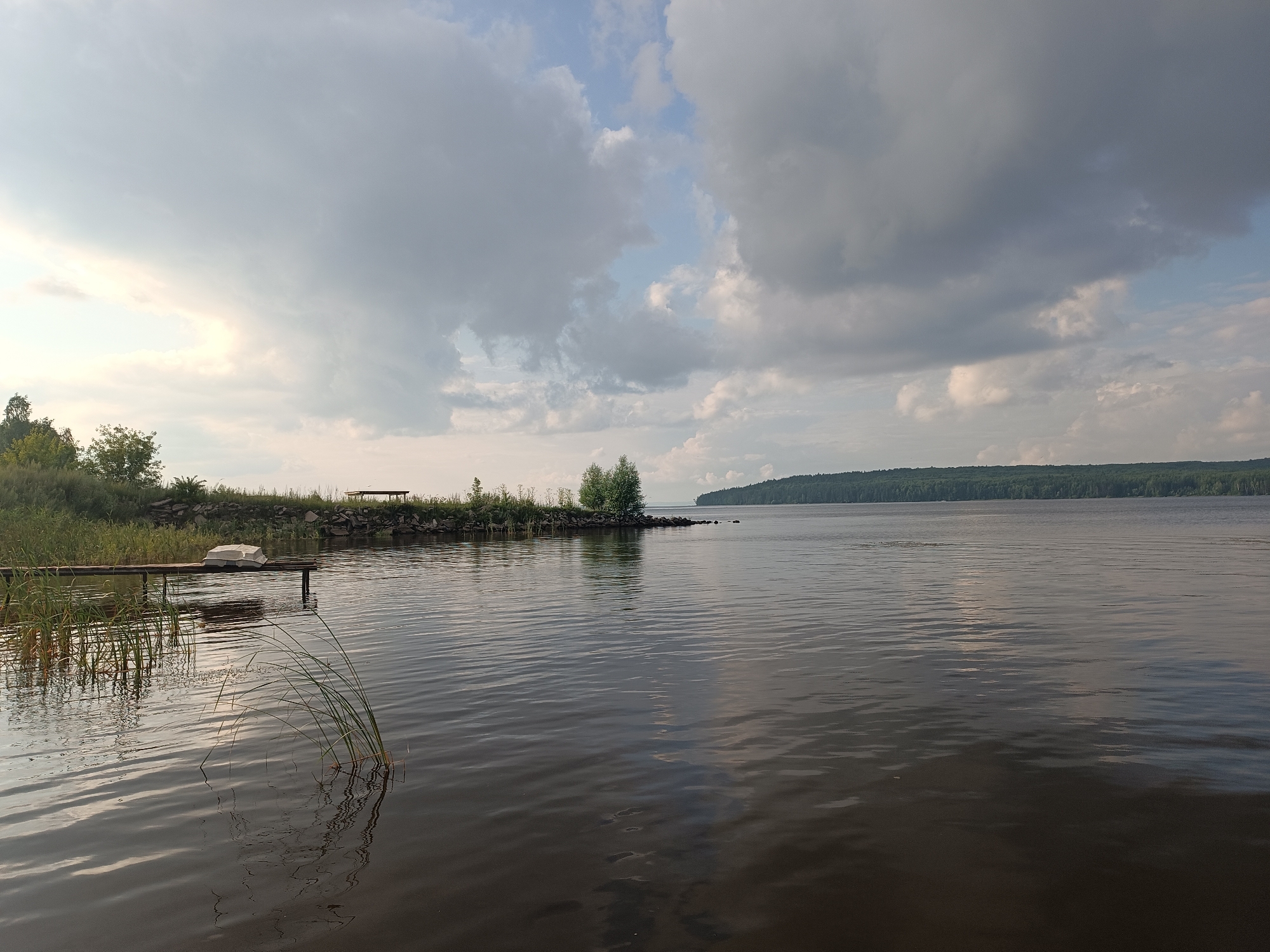 The image depicts a wide expanse of calm water, likely a river or lake, with gentle ripples reflecting the overcast sky. On the left bank, there is a wooden dock extending into the water, with a small white boat resting on it. Reeds and tall grass grow along the bank. Further along the bank, a small cluster of trees is visible, as well as a rocky shoreline. In the distance, the opposite bank of the water features a dense line of trees stretching across the horizon under a dramatic sky filled with various types of clouds, from light and wispy to dark and heavy.
This scene is consistent with views of the Kama River in the city of Chaikovsky, Russia, specifically near its shores, where such natural landscapes with docks and boats are common. There are no people or animals visible in the image. The overall atmosphere is serene and natural.