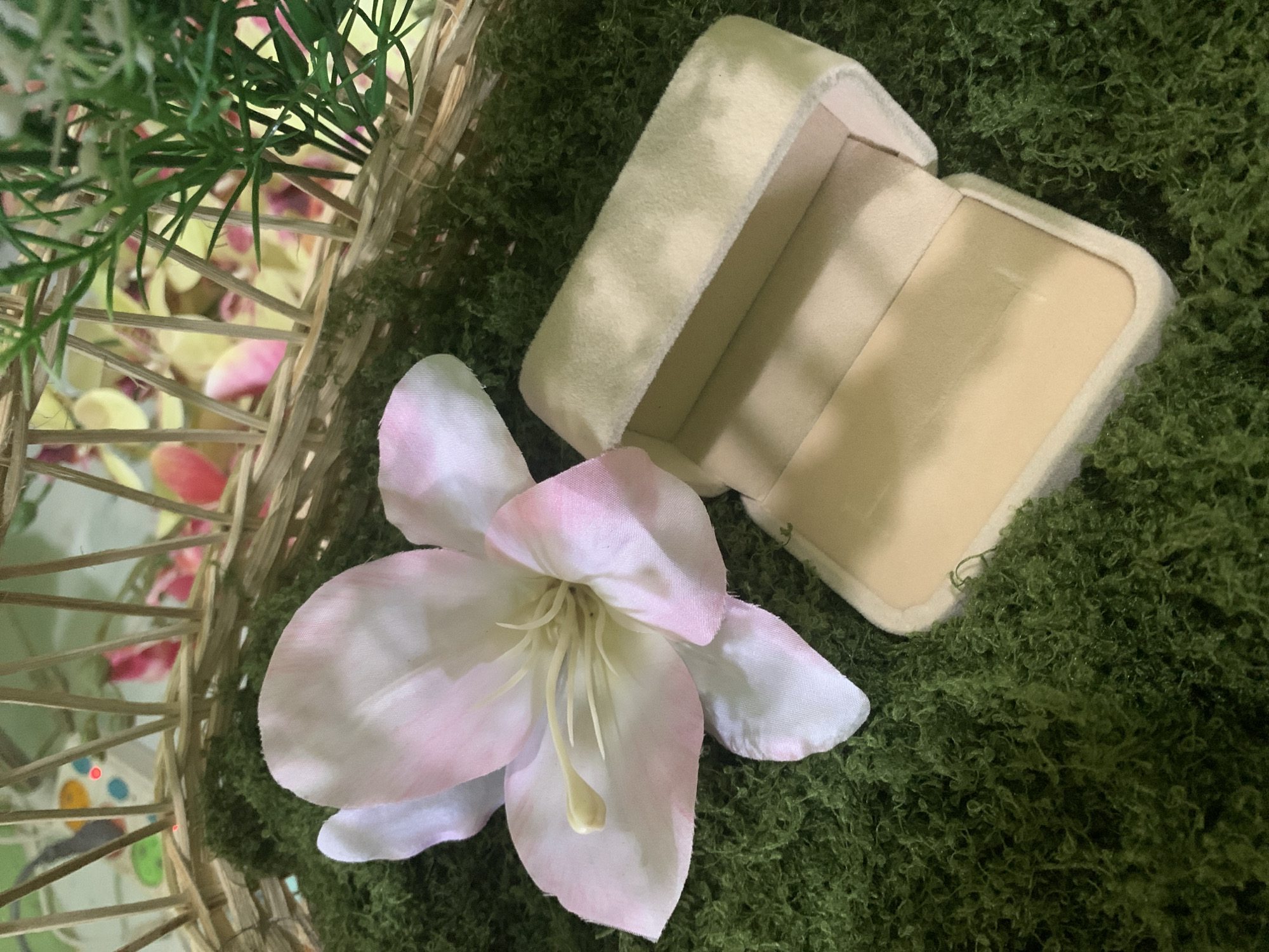A top-down, close-up shot shows a light pink and white flower with delicate petals and yellow stamens resting on a bed of green moss. To the right of the flower is an open, beige velvet ring box, also on the moss. In the background, a portion of a woven basket is visible, containing some greenery and blurred pink and yellow elements. The scene appears to be staged, possibly for a product photograph, and has no discernible connection to Kuala Pembuang, Indonesia.