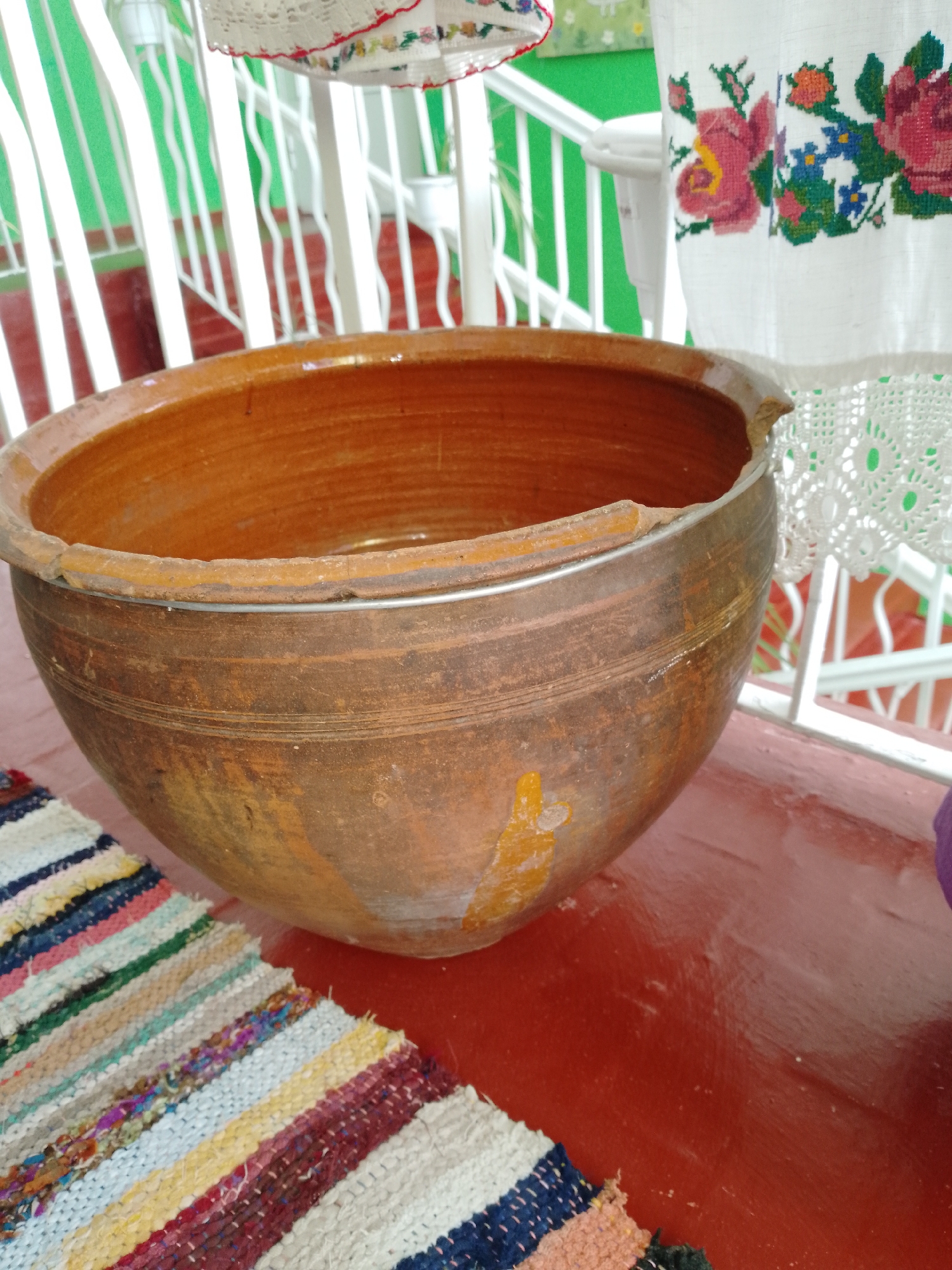 The image captures a static, brightly lit scene centered around a large, rustic, reddish-brown clay pot. The pot, positioned prominently in the foreground, shows signs of age and wear with varying shades of brown, some chipped areas on its rim, and a metallic band encircling its upper section, suggesting reinforcement or repair. Its surface has visible striations and some lighter, possibly stained or worn, patches.
The pot rests on a reddish-brown floor, which appears to be painted wood or concrete. In the bottom left corner, a colorful striped woven mat or rug is partially visible, made from various fabric scraps in hues of white, blue, red, yellow, green, and pink.
In the background, on the right side, a white fabric with vibrant, intricate floral embroidery hangs. The embroidery features large red roses, smaller blue flowers, and green leaves, with a delicate white crocheted lace trim along its bottom edge. Behind this fabric and the pot, white vertical railings or balusters are visible, set against a bright, uniform green background, possibly a wall or lush foliage. The railings create a sense of depth and suggest an outdoor or semi-outdoor setting like a porch or balcony.
No people are present, and no specific activity is taking place; the scene appears to be a display of traditional household items. The overall impression evokes a sense of cultural heritage and rustic charm, typical of a home or folk art exhibit in a rural Ukrainian setting like Ustyvytsia. There is no visible text in the image.