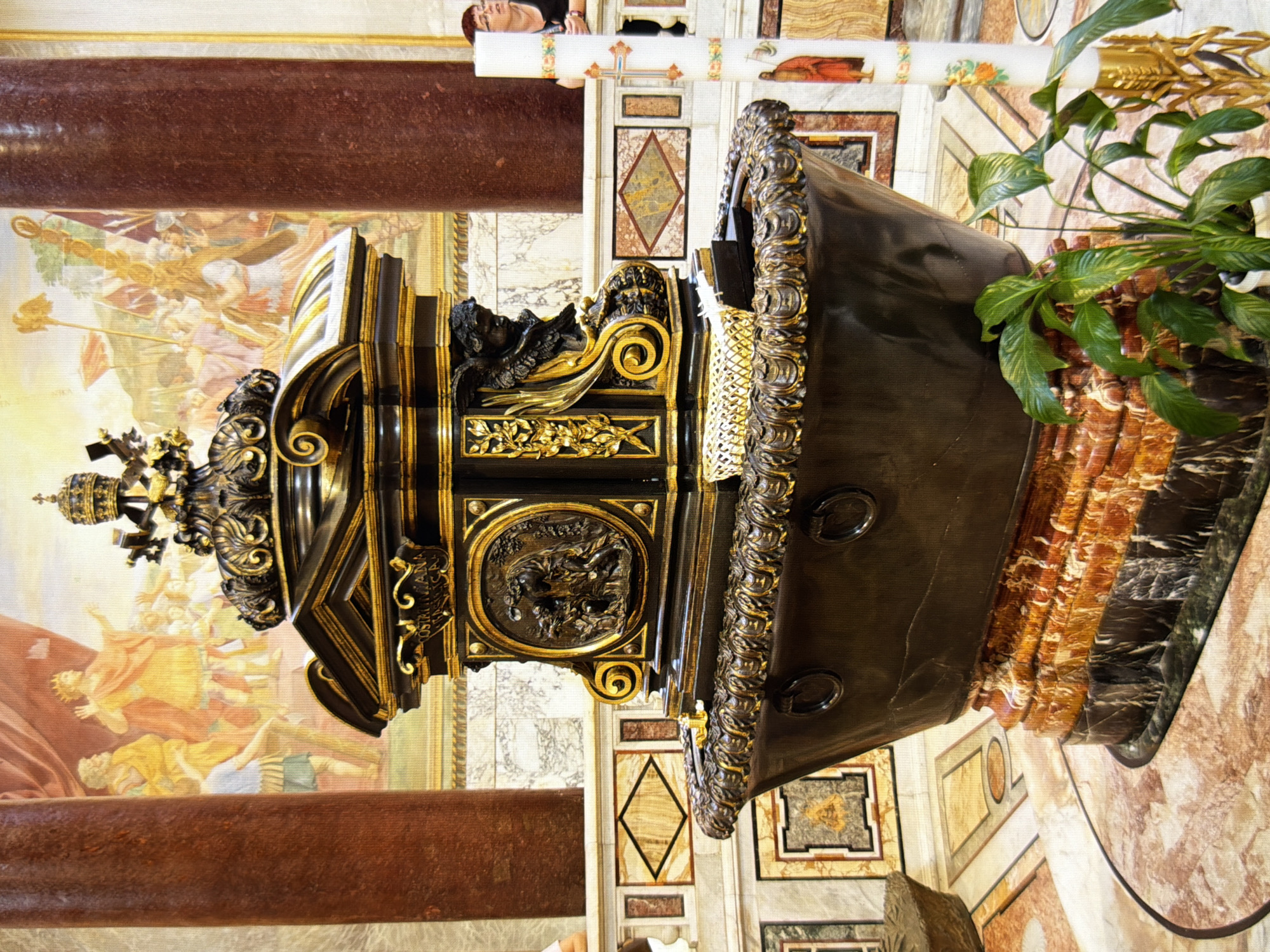The image shows a close-up of a large, ornate, dark bronze baptismal font, likely made of metal and marble, with intricate gold detailing. The font is filled with water, and a small white basket with white objects sits inside. The font rests on a multi-layered, ornate base constructed from what appears to be a dark, rough stone at the bottom and several layers of polished, veined marble with shades of orange and white on top.
The background features a mural with depictions of figures in historical or religious attire, painted in warm, muted colors. Two large, dark red marble columns flank the font. To the right of the font, a tall, white Paschal candle is visible, decorated with colorful bands and images, including a cross at the top and a figure of a robed man near the bottom. A woman with glasses and dark hair is partially visible behind the candle, leaning on a balustrade. In front of the font, a healthy green potted plant with large, glossy leaves is situated. The floor is a patterned marble mosaic in shades of beige, orange, and pink. The environment appears to be an interior religious space, possibly a church or cathedral, with a grand and ceremonial atmosphere.
There is no indication in the image that this scene is located in Kyiv, Ukraine. The architectural style and the artwork depicted are not specific to any particular location in Kyiv.