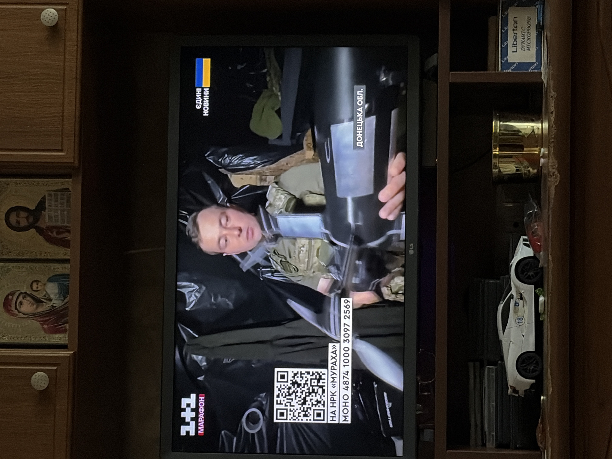 The image shows a television screen displaying a news broadcast from Ukraine. A man in military attire is holding a large cylindrical object, possibly a weapon or piece of equipment. The broadcast includes Ukrainian text overlays indicating "1+1 Marathon," "News," and "Donetsk Oblast." To the left of the screen, a QR code and some numbers are displayed. Above the television, on a wooden shelf, are two religious icons. Below the television, on a lower shelf, a white toy sports car is visible, along with other miscellaneous items including a gold-colored container and a box labeled "Liberton DYNAMIC MICROPHONE." The overall environment appears to be a domestic setting.