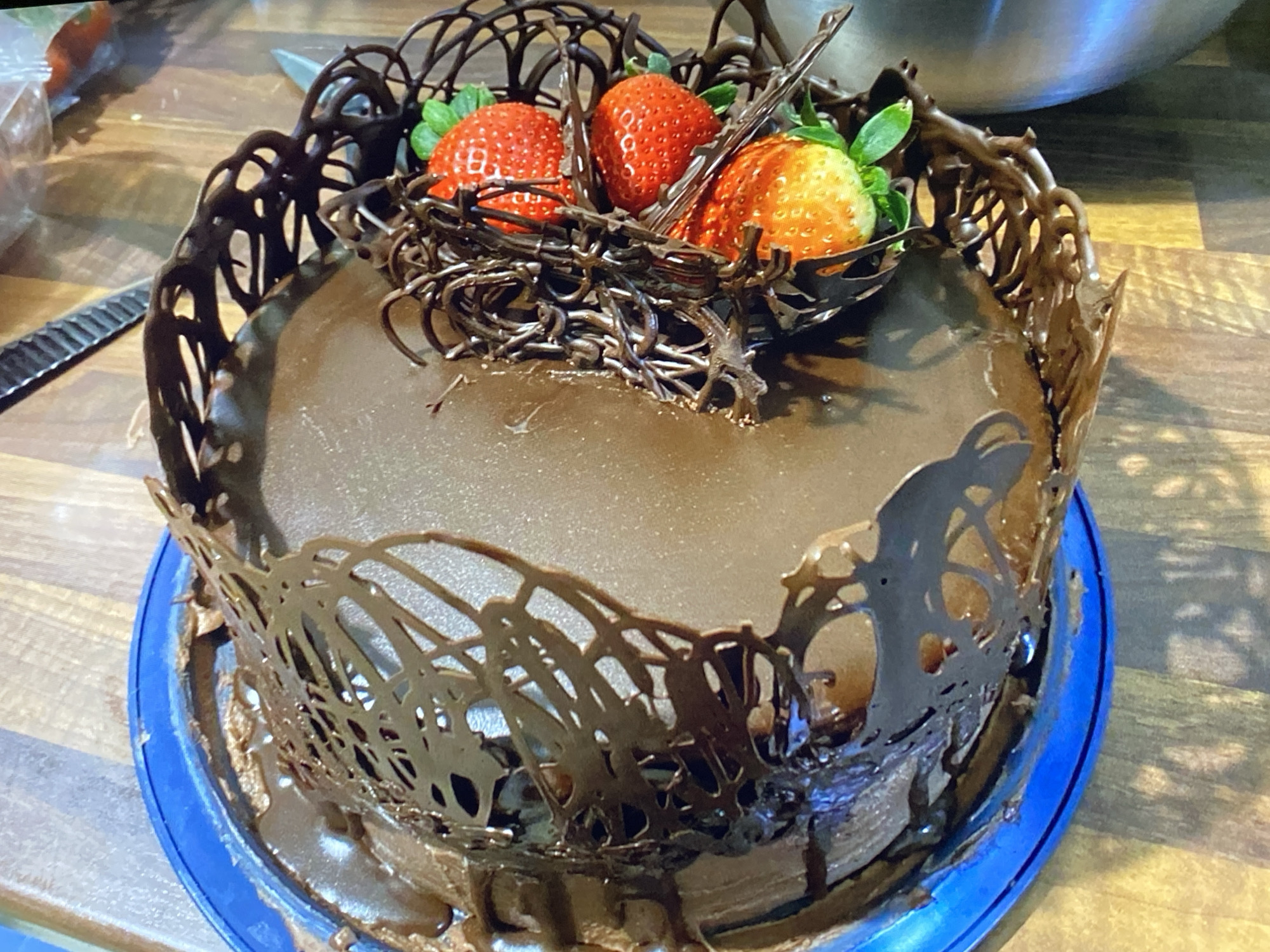 A close-up shot shows a decadent chocolate cake decorated with elaborate chocolate lace around its sides and a chocolate nest filled with three strawberries on top. The cake is covered in a smooth, rich chocolate frosting, and the chocolate lace creates an intricate, openwork design. The strawberries, with their vibrant red skin and green leaves, add a fresh contrast to the dark chocolate. The cake sits on a bright blue plate, which rests on a wooden surface that appears to be a countertop or table. In the background, a stainless steel bowl is partially visible, suggesting a kitchen environment. The lighting is warm and natural, indicating it might be daytime. There is no visible text in the image.