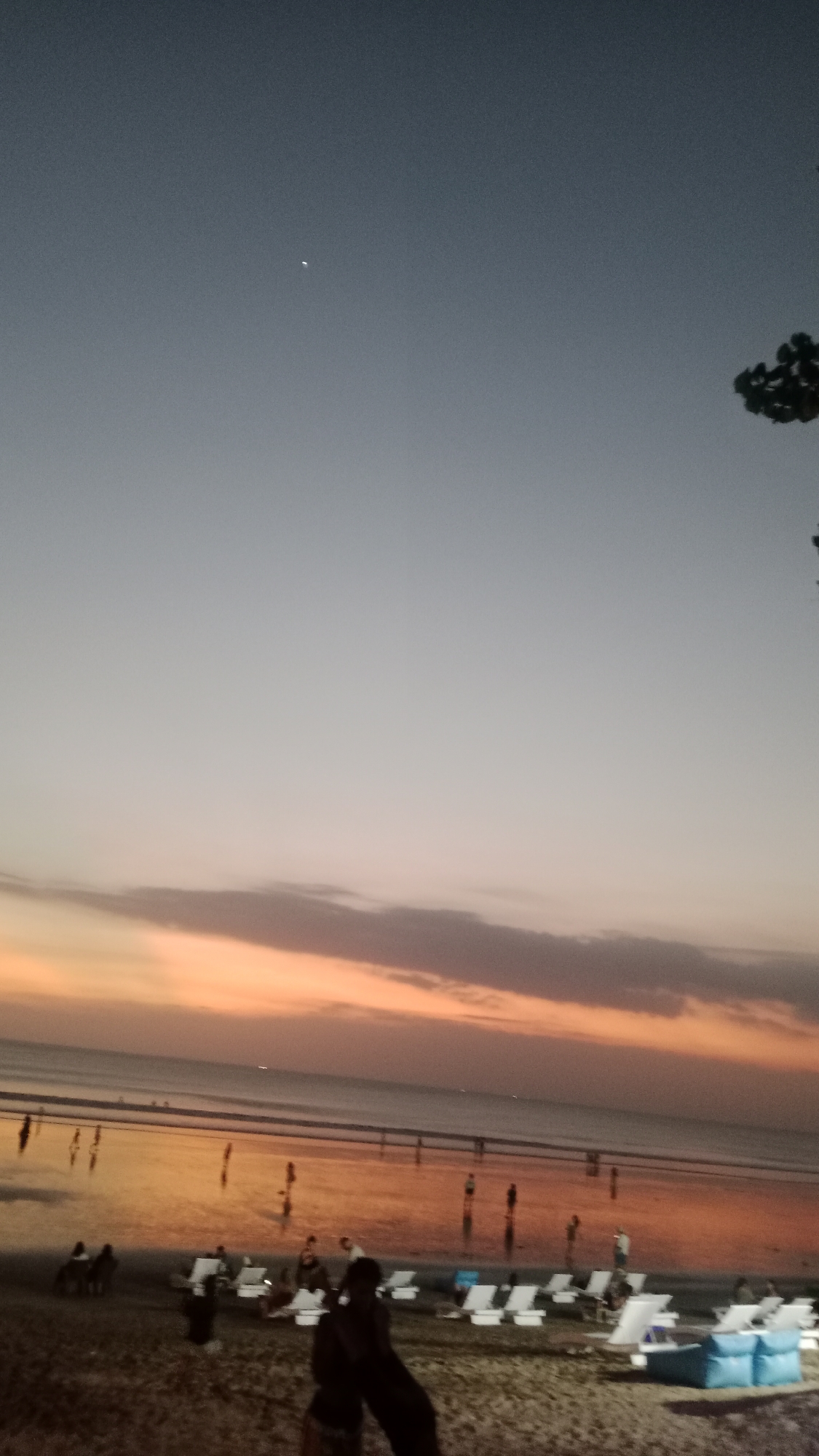 This is a photograph taken at dusk on Kuta Beach, Bali, Indonesia. The sky displays a gradient of colors, from a deep blue at the top to soft orange and pink hues closer to the horizon, indicating the setting sun. A faint crescent moon is visible in the upper portion of the sky.
The beach itself is sandy, with scattered lounge chairs and some people relaxing or walking. In the middle ground, the ocean is calm and reflects the colors of the sunset. Numerous people are visible along the shoreline, some standing in the shallow water and others walking on the wet sand. Their figures are silhouetted against the bright water and sky.
In the foreground, more lounge chairs are arranged, and several individuals are present, some seated and others standing. The lighting suggests it is twilight, and artificial lights from nearby establishments may be contributing to the illumination. The overall scene captures a peaceful evening atmosphere on a popular tourist beach.