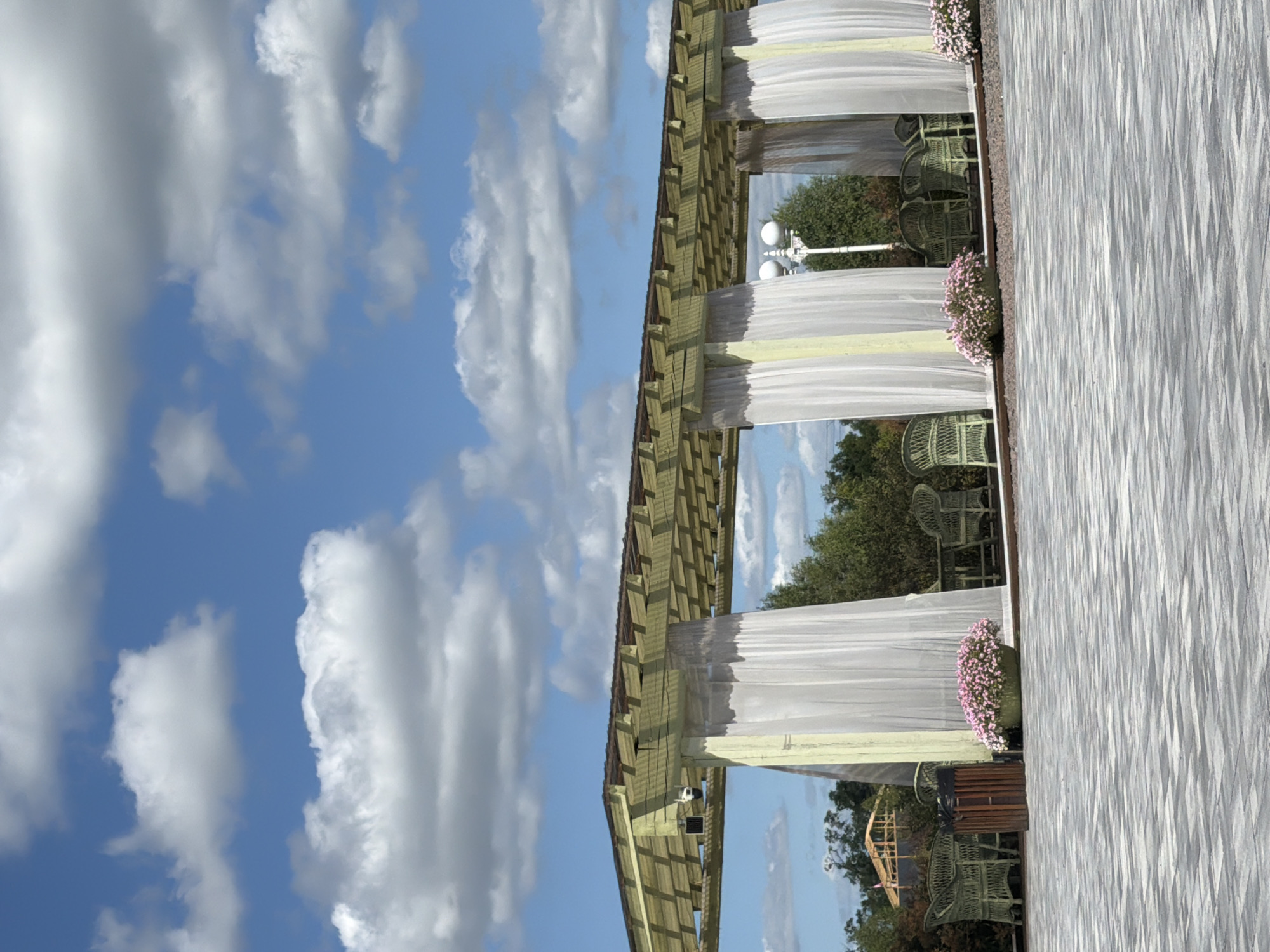 The image shows an outdoor seating area under a pergola in Severynivka, Ukraine. The pergola is constructed of light-colored wood and has a slatted roof. White curtains hang from the sides of the pergola, creating separate seating alcoves. Inside the alcoves, there are several green wicker chairs and tables. Two planters filled with pink flowers are visible on the ground. The ground itself is paved with a patterned grey and white stone. In the background, there are trees and a partially constructed wooden structure, possibly a building or stage. The sky above is blue with scattered white clouds. A street lamp with two globes is visible between two of the pergola's support columns. The scene appears to be a tranquil outdoor dining or event space.