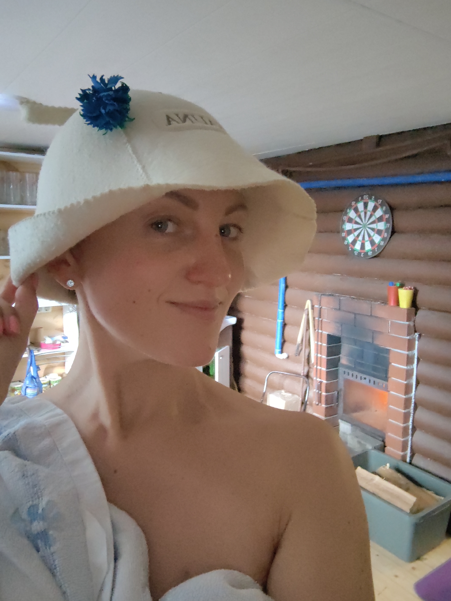 A woman with blonde hair is standing in what appears to be a sauna or rustic room. She is wearing a white felt hat decorated with a blue pom-pom and an earring. Her left shoulder is draped with a white towel. In the background, a dartboard is mounted on a wooden wall above a brick fireplace with a fire burning. There are also logs in a green container next to the fireplace, and shelves with various items are visible to the left. The room has a ceiling with visible lights. The overall impression is one of a leisure or relaxation setting. The location is identified as Фаніпальскі сельскі Савет, Belarus.