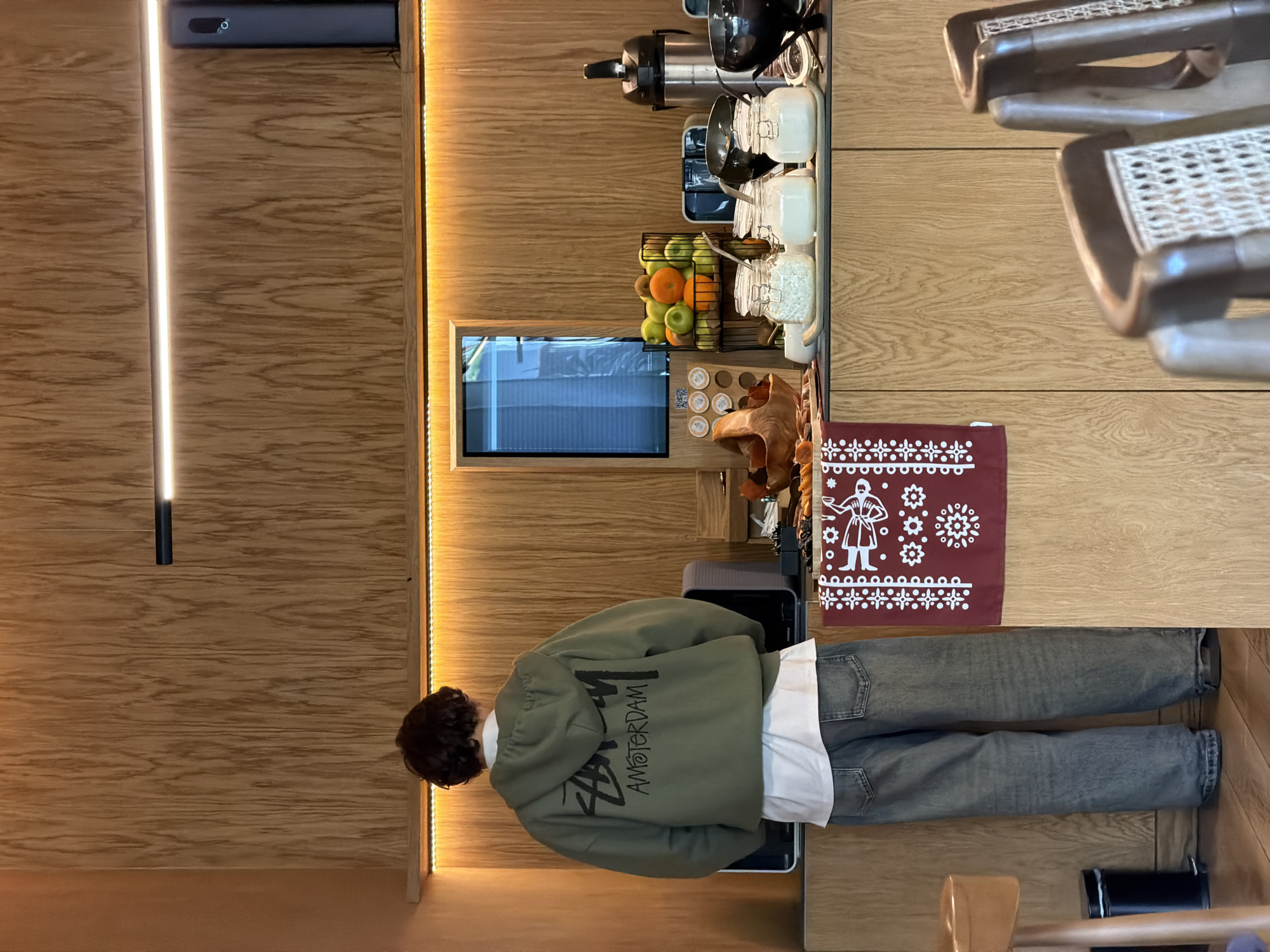 The image shows a person standing in a cafe or restaurant in Tbilisi, Georgia. The person is wearing a green hoodie with "Amsterdam" written on the back, a white shirt, and gray jeans. They are standing near a coffee station, which features a coffee maker, jars of toppings, a fruit basket, and a small screen. The establishment has wood paneling on the walls and furniture, and the lighting appears to be warm and inviting. There are chairs visible to the right. A burgundy napkin is positioned beside the coffee station.