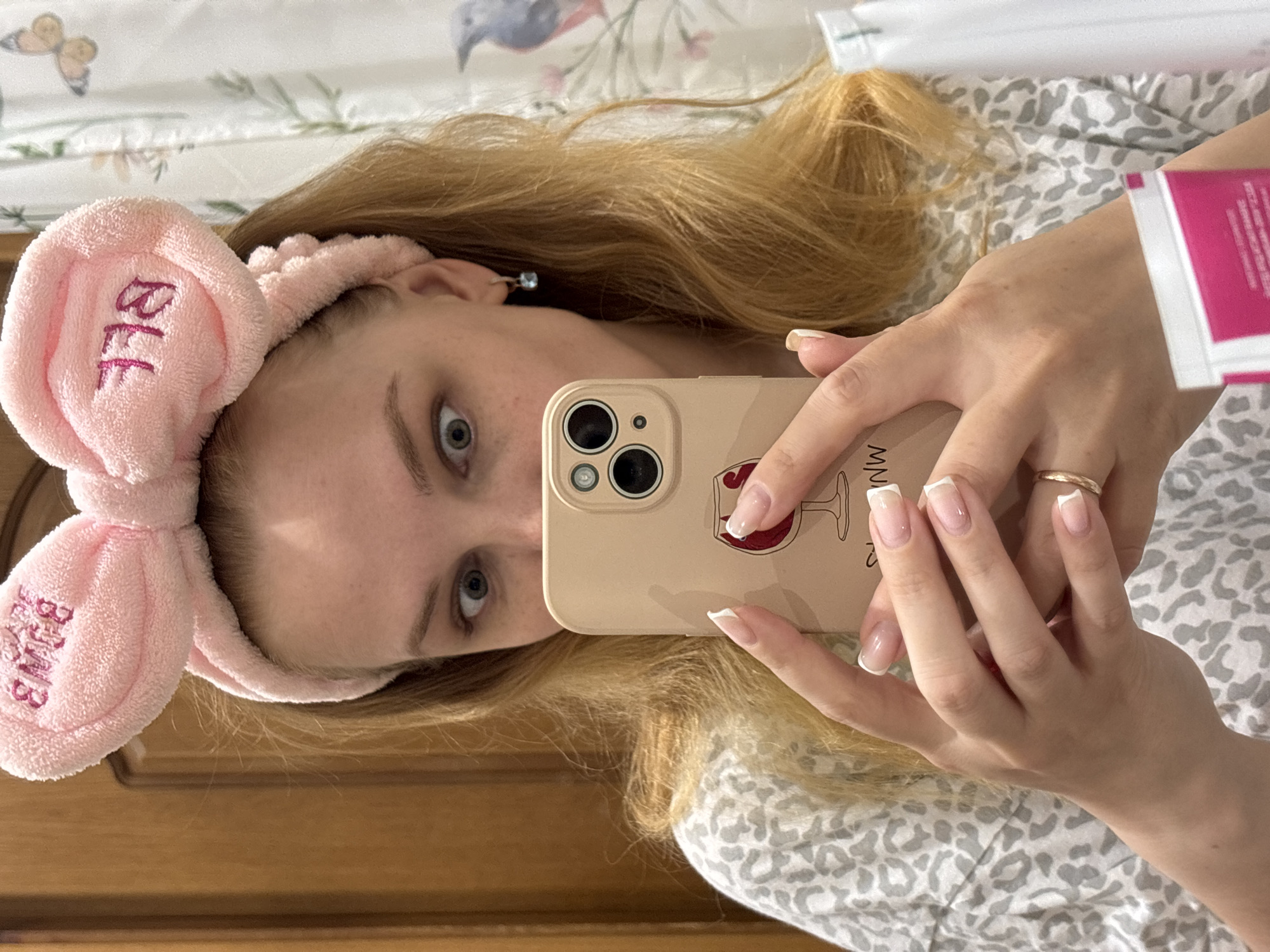 A young, fair-skinned woman is holding an iPhone with a wine glass design on the case, taking a selfie. She is wearing a pink headband with the letters "BFF" embroidered on it. She is wearing a gold ring on her ring finger. The woman is lying on a bed with a gray and white patterned sheet. The background includes a white curtain with floral patterns. Rusanovo, Russia is not visible.